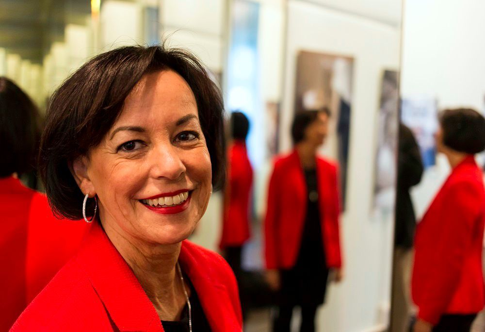 Porträtaufnahme Jutta Törnig-Struck, die in die Kamera lächelt. Verschwommen im Hintergrund ist sie mehrfach gespiegelt in Ganzkörperansicht zu sehen, da sie mit dem Rücken zu einem Spiegel steht der sich wiederum in mehreren Bilderrahmen widerspiegelt.