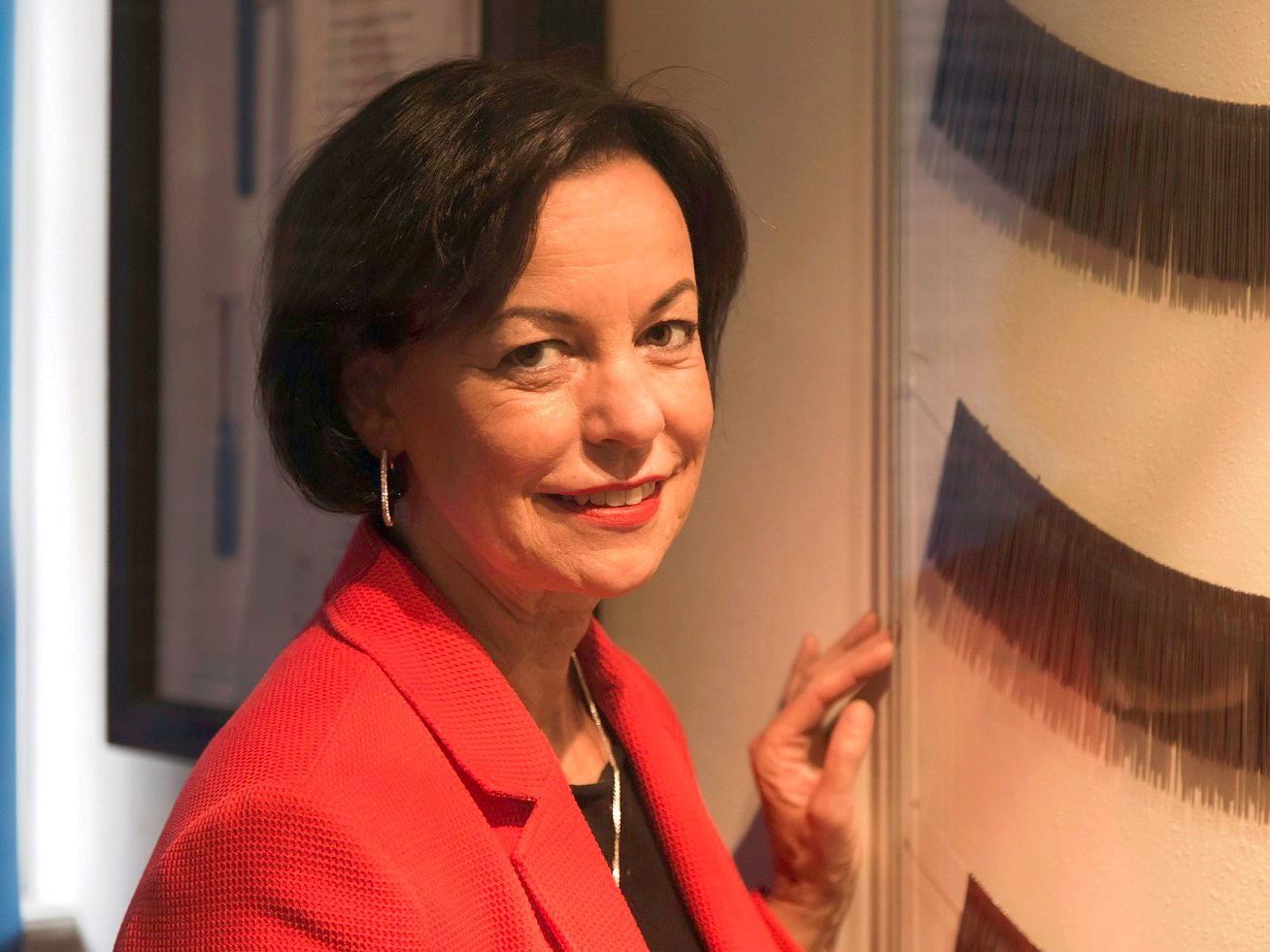 Jutta Törnig-Struck steht im Museum vor einem flachen Glasrahmen an der Wand, hinter dem alte Nadeln zu sehen sind. Die Nadeln sind wie Colliers aufgereiht.