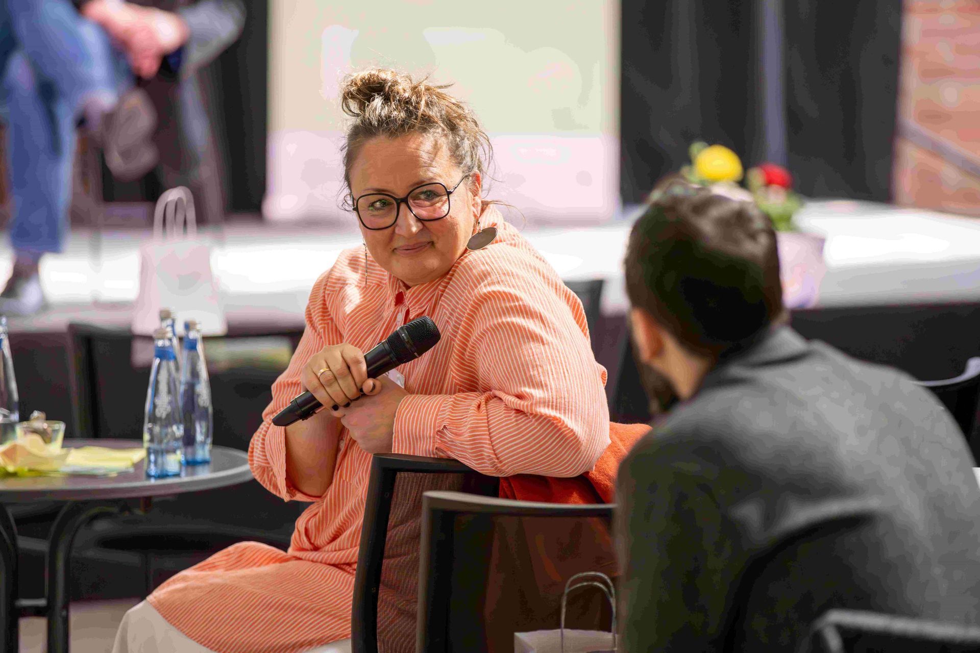 Eine Teilnehmerin sitzt auf einem Stuhl mit dem Mikrofon in der Hand und dreht sich zuhörend dem Moderator zu.