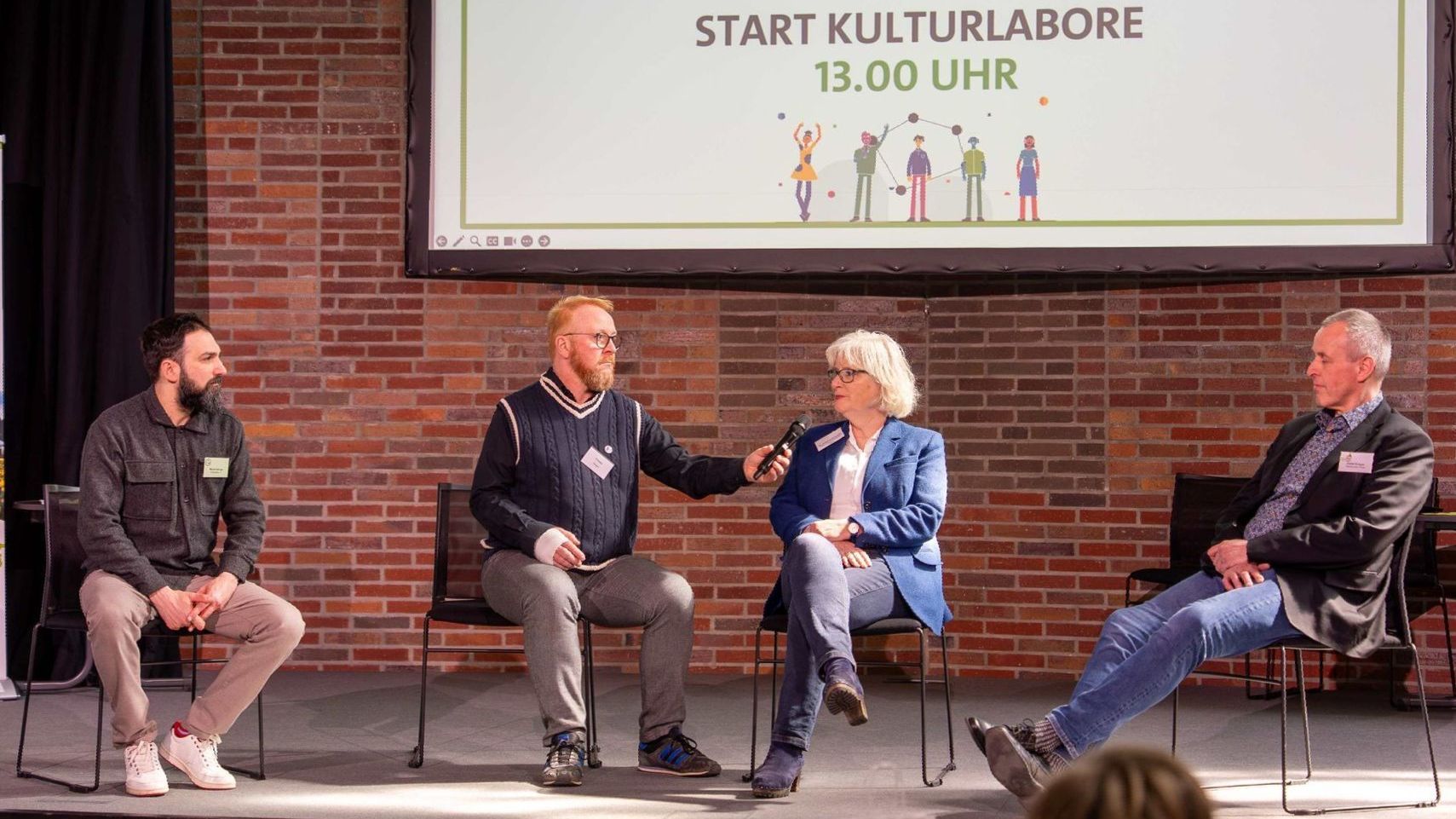 Vier Teilnehmende des Kulturlabors 2 sitzen auf Stühlen auf der Bühne. Zwei sind in einer Interview miteinander, die beiden anderen hören zu.
