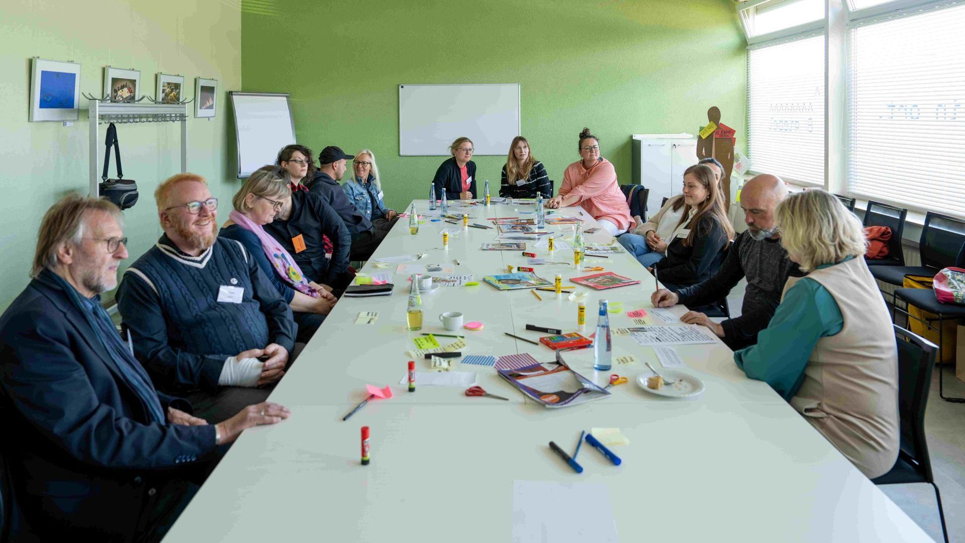 13 Teilnehmende des Kulturlabor 3 sitzen an einem langen Tisch sich gegenüber. Auf dem Tisch liegen verstreut viele bunte Zettel, Stifte, Klebestifte und Zeitschriften.
