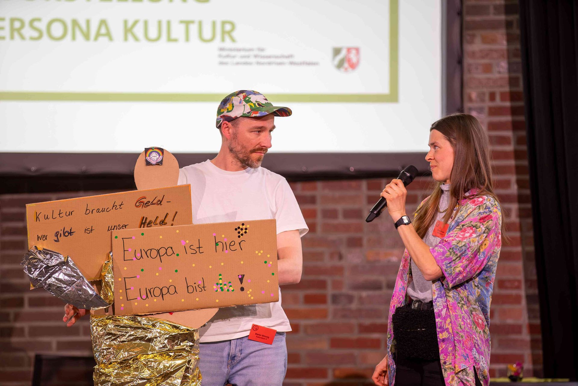 Die Leiter:innen des Kulturlabors 4 Anne von Deckert und Stefan Bünnig stehen mit ihrer Kulturpersonal auf der Bühne. Anne von Deckert hält ein Mikrofon in der Hand und spricht zu Stefan Bünnig.