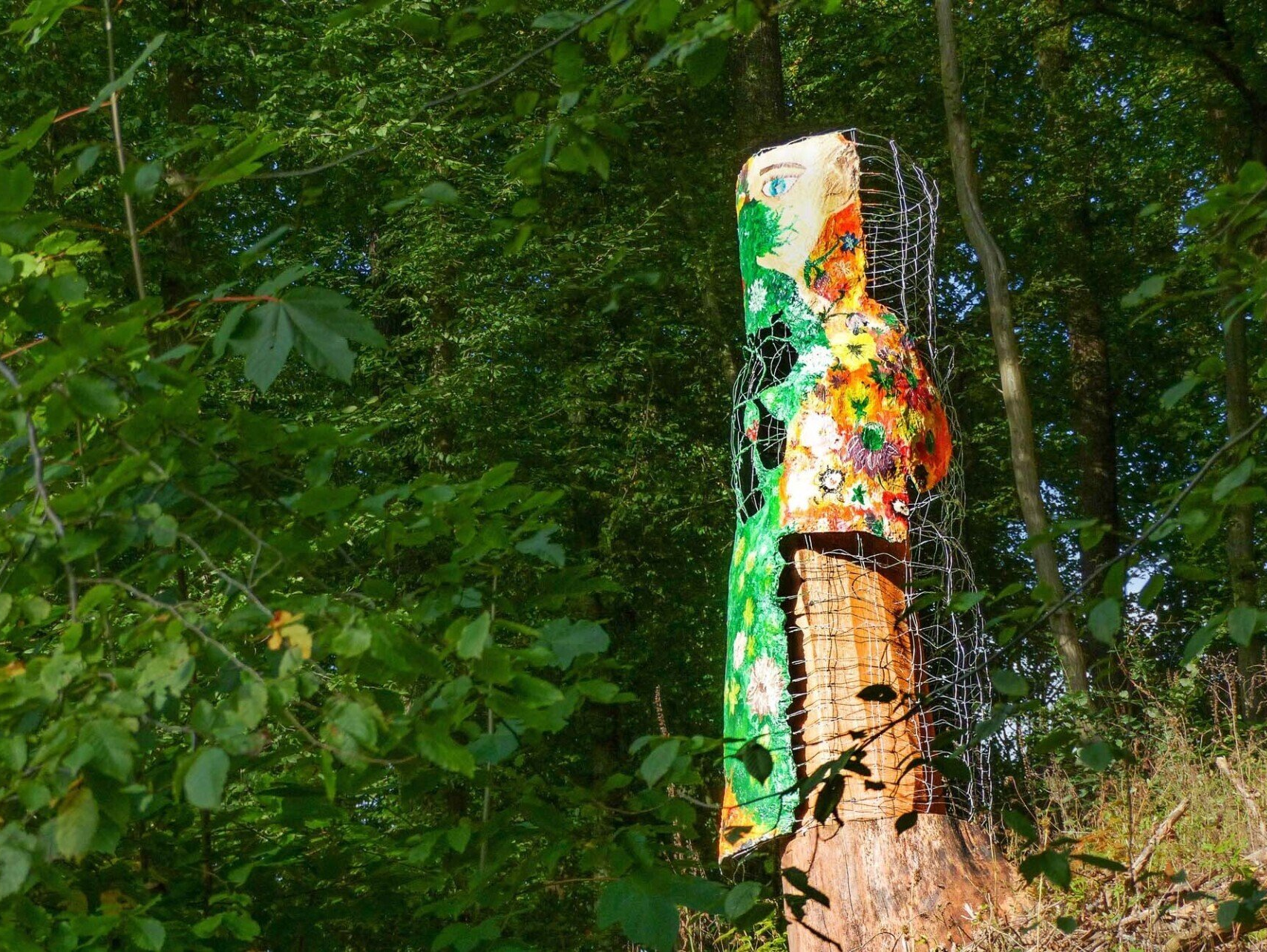 Ein Kunstwerk steht an einem Hang im Grünen. Das Kunstwerk besteht aus einem Rest eines aufragenden Baumstamms mit angemalter Rinde verkleidet.