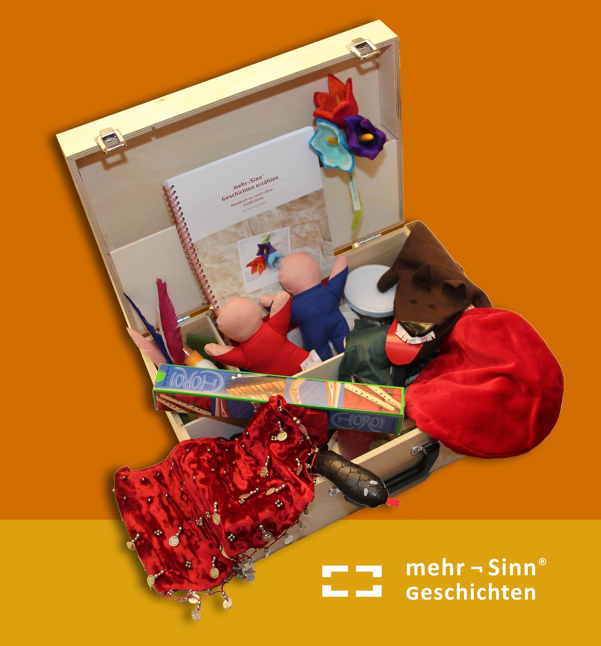 Geöffneter Mehrsinn-Erzählkoffer mit Requisiten wie Handpuppen, Stoffe, Büch, Kissen u.a. darunter die Schriftmarke mehr-Sinn Geschichten