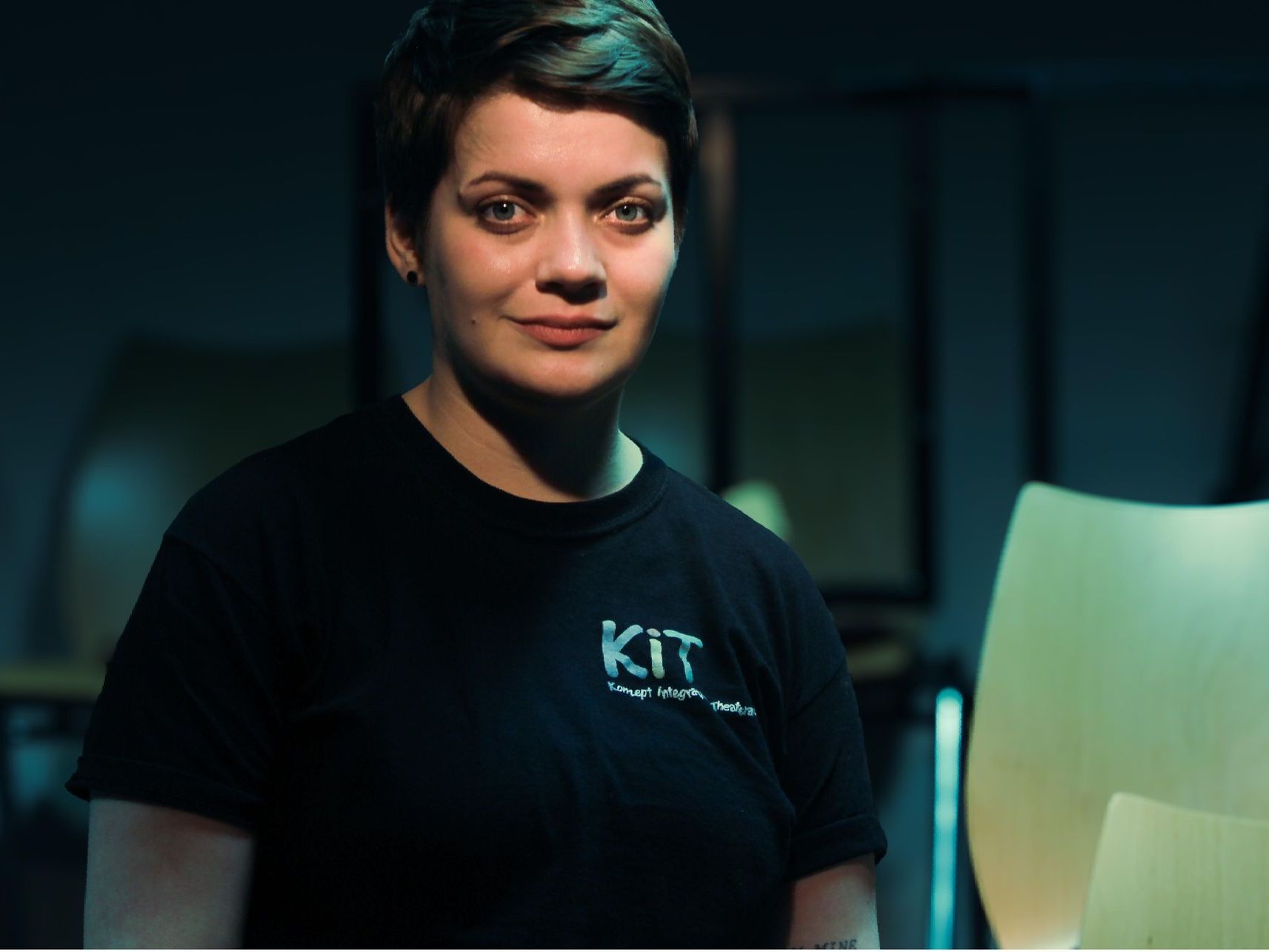 Porträt von Lea Schnalke, schwarzes T-Shirt mit Schriftzug KIT - Konzept integrative Theaterarbeit, kurze dunkle Haare. Im Hintergrund sieht man die Sitzreihen der Bühne der Integrativen Theaterwerksatt.