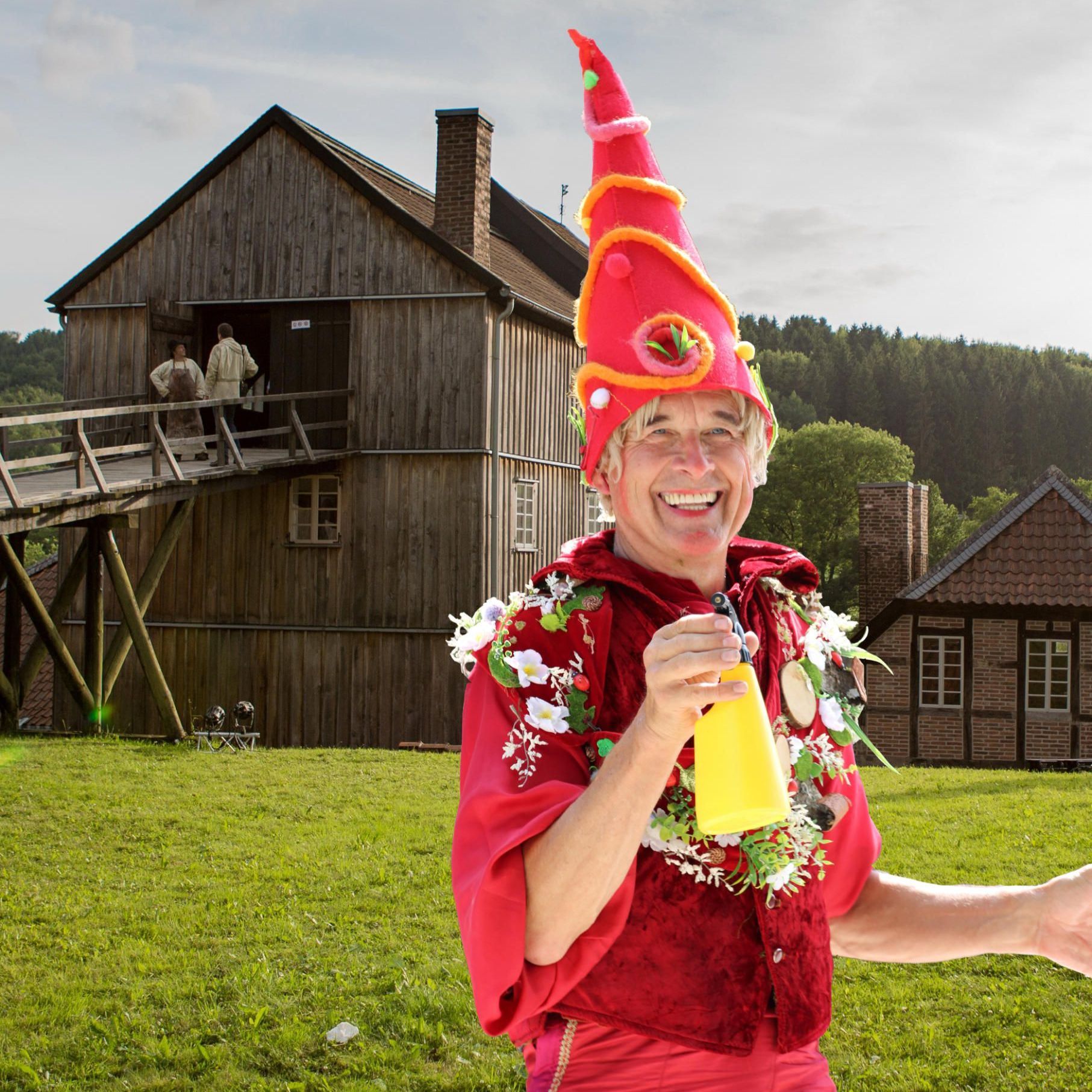 Mann als Zwerg verkleidet mit roter spitzer Zwergenmütze, roter Kleidung hält eine Spritzpistole in der Hand und steht vor der Luisenhütte.