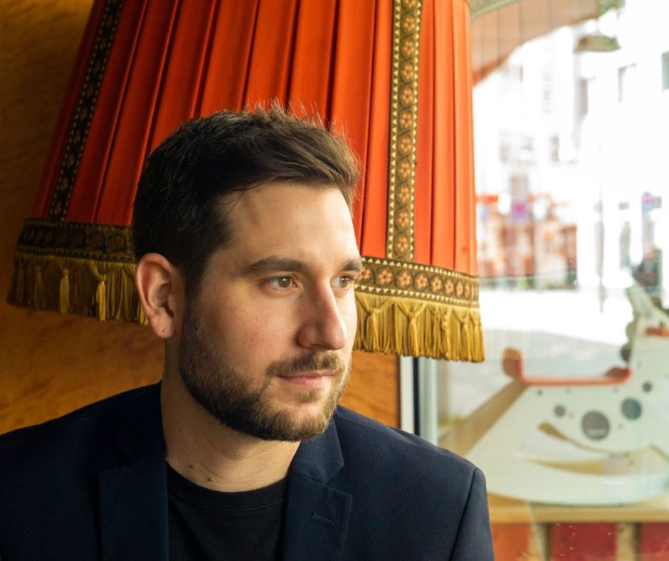 Porträtaufnahme von Marc Debie, vor einem alten roten Lampenschirm sitzend. Er schaut seitlich aus dem Schaufenster der Theaterbotschaft auf die Hagener Einkaufsstraße