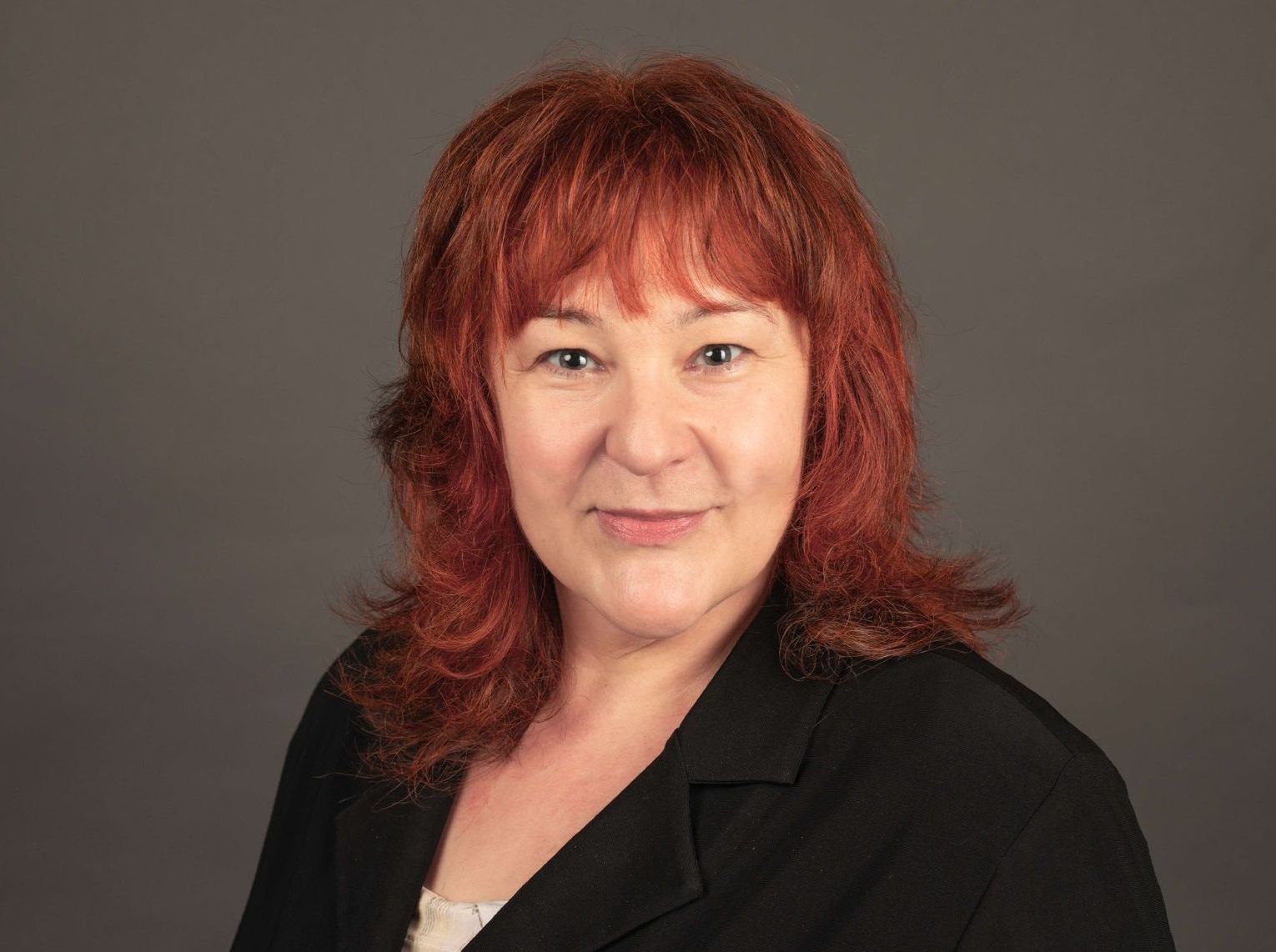 Frau Redlberger schaut leicht lächelnd in die Kamera. Die rötlich halblangen Haare heben sich vom grauen Hintergrund ab.