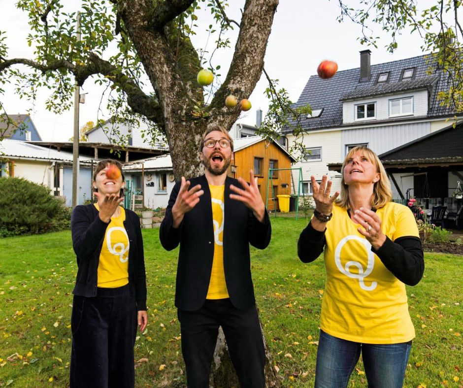 Drei Mitglieder der QulturwerkstattJjonglieren im Garten des Kulturzentrums vor einem alten Apfelbaum mit Äpfeln. Alle sind mit gelben T-Shirts bekleidet, darauf ist das Logo Q zu sehen.