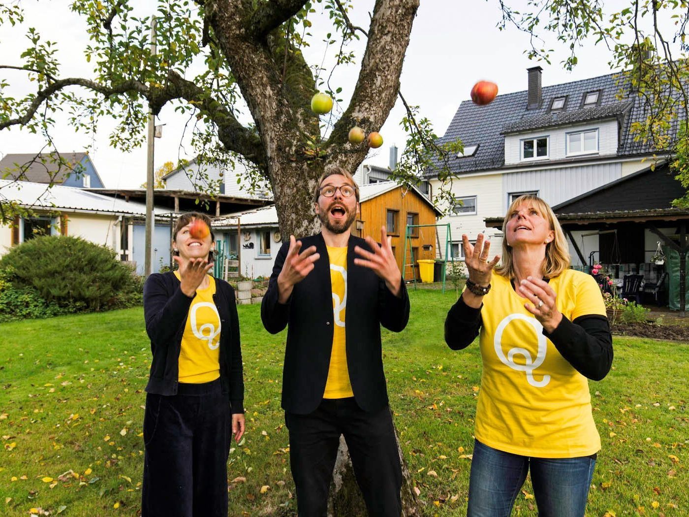 Drei Mitglieder der QulturwerkstattJjonglieren im Garten des Kulturzentrums vor einem alten Apfelbaum mit Äpfeln. Alle sind mit gelben T-Shirts bekleidet, darauf ist das Logo Q zu sehen.