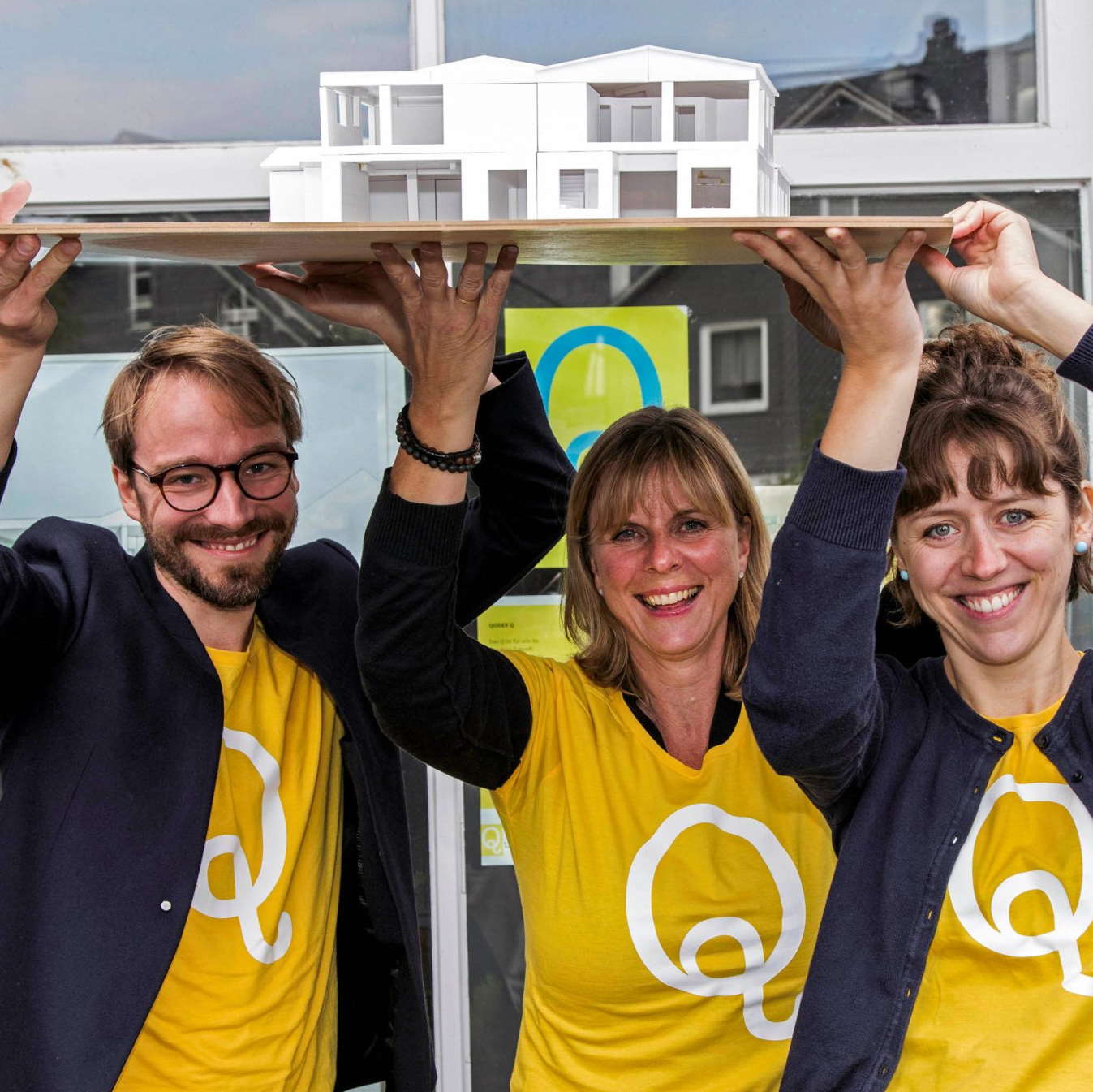 Drei Mitglieder der Qulturwerkstatt tragen ein Modell des Gebäudes über ihren Köpfen. Alle sind mit einem gelben T-Shirt mit dem Q für Qulturwerkstatt bekleidet.