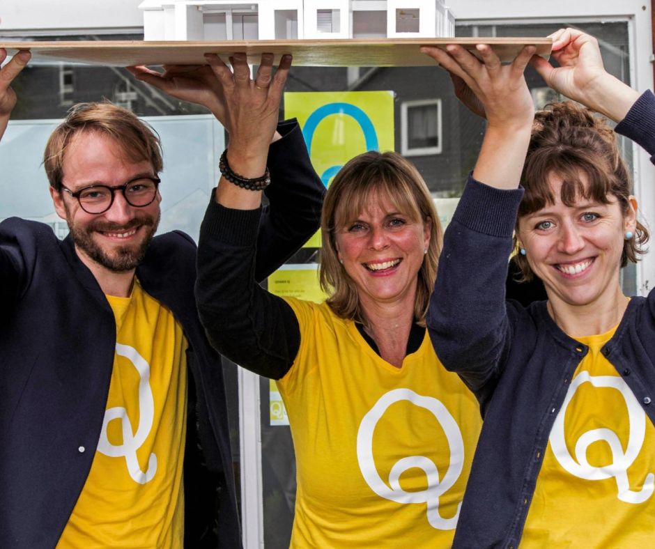 Drei Mitglieder der Qulturwerkstatt halten das Modell der geplanten Kulturzentrums über ihren Köpfen. Alle sind mit gelben T-Shirts bekleidet, darauf ist das Logo Q zu sehen.