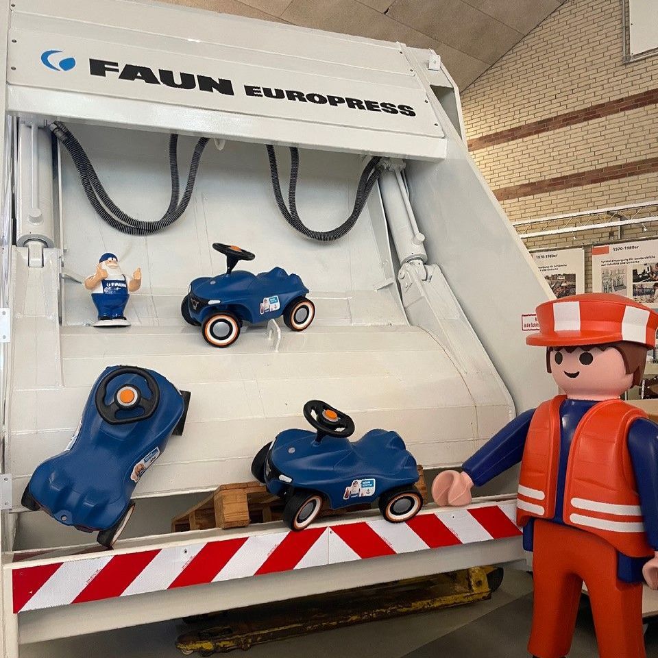 Lebensgroße Playmobilfigur steht vor dem Einwurf eines Müllwagens, auf dem Bobbycars trapiert sind.