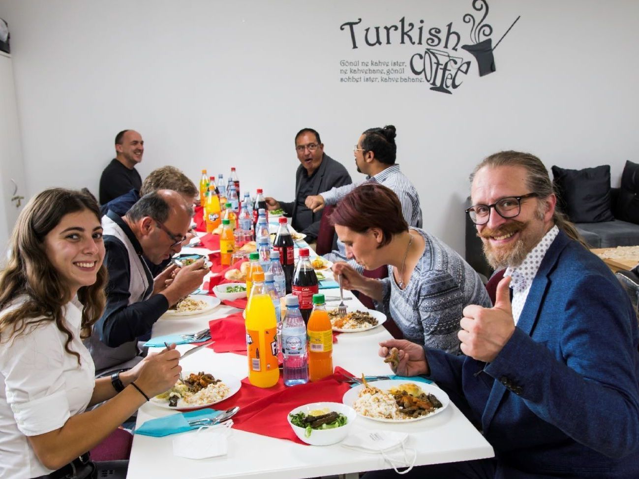 Menschen verschiedener Nationen sitzen beim gemeinsamen Essen an einem langen Tisch in einem Raum der Attendorner Moschee. Alle sind ins Essen und ins Gespräch vertieft, der Mann im Bildvordergrund reckt den Daumen in die Höhe und lacht.