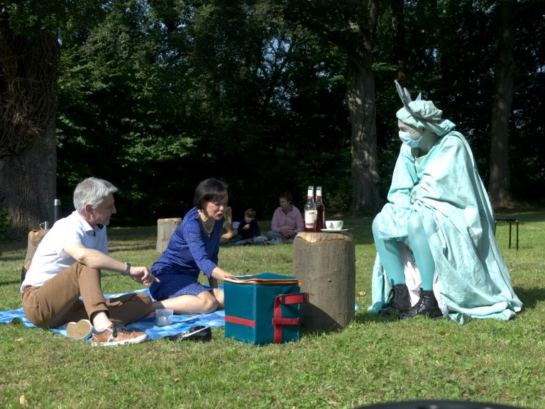 Ein Mann und eine Frau sitzen auf einer Decke in einem Park und machen Picknick. Daneben sitzt eine als New Yorker Freiheitsstatue verkleiderte Person.
