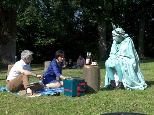 Zwei Menschen sitzen auf einer Picknickdecke in einem Park. Daneben sitzt auf einem Stuhl einer als New Yorker Freiheitsstatue verkleideter Walk-Act-Künstler.