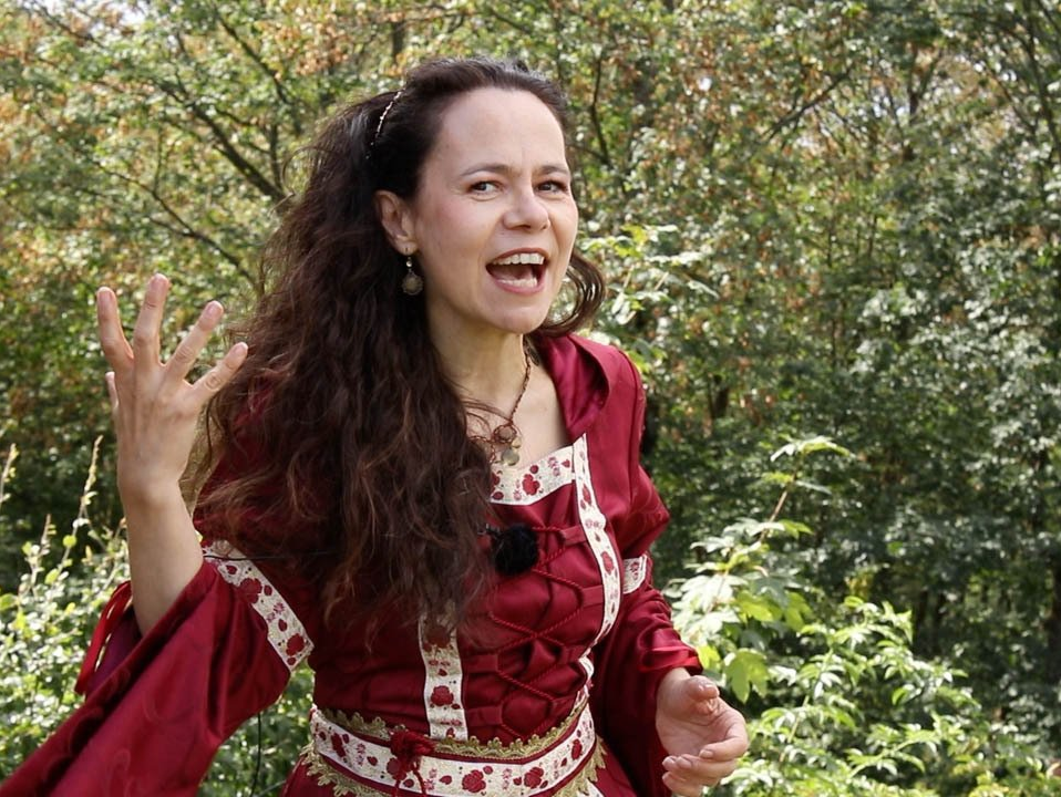 Eine Frau mit langen Jahren und in mittelalterlicher Gewandung erzählt mit vollem Körpereinsatz eine Geschichte. Das Foto wurde in einem lichten Wald aufgenommen.