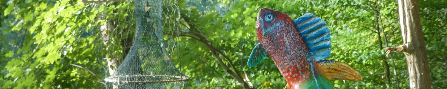 Ein bunter Fisch ragt als Kunstwerk an einem Flußufer übergroß empor, neben ihm hängt eine Fischreuße im Baum.