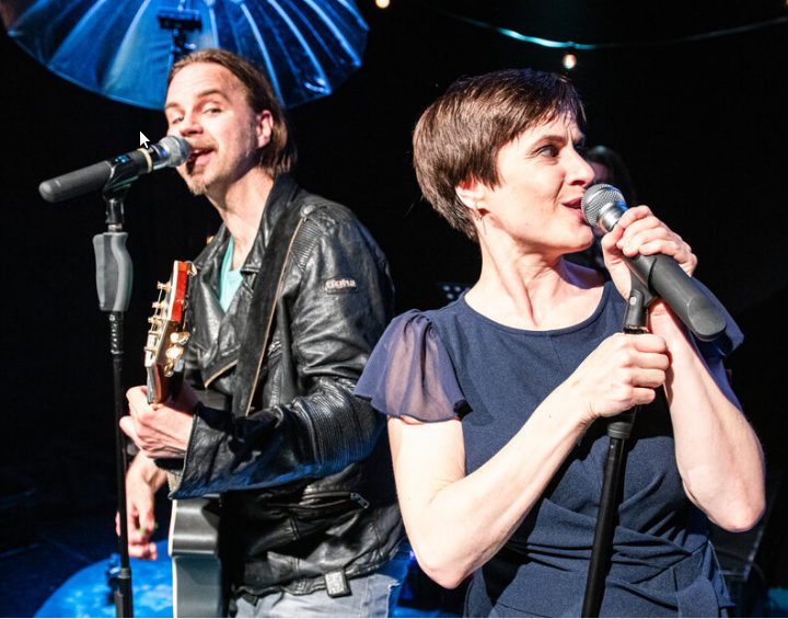 Die Hauptdarsteller Milan Pesl und Irna Ries auf der Bühne. Ina Ries singt in ein Mikrofon, Milan Pesl singt und spiel Gitarre.