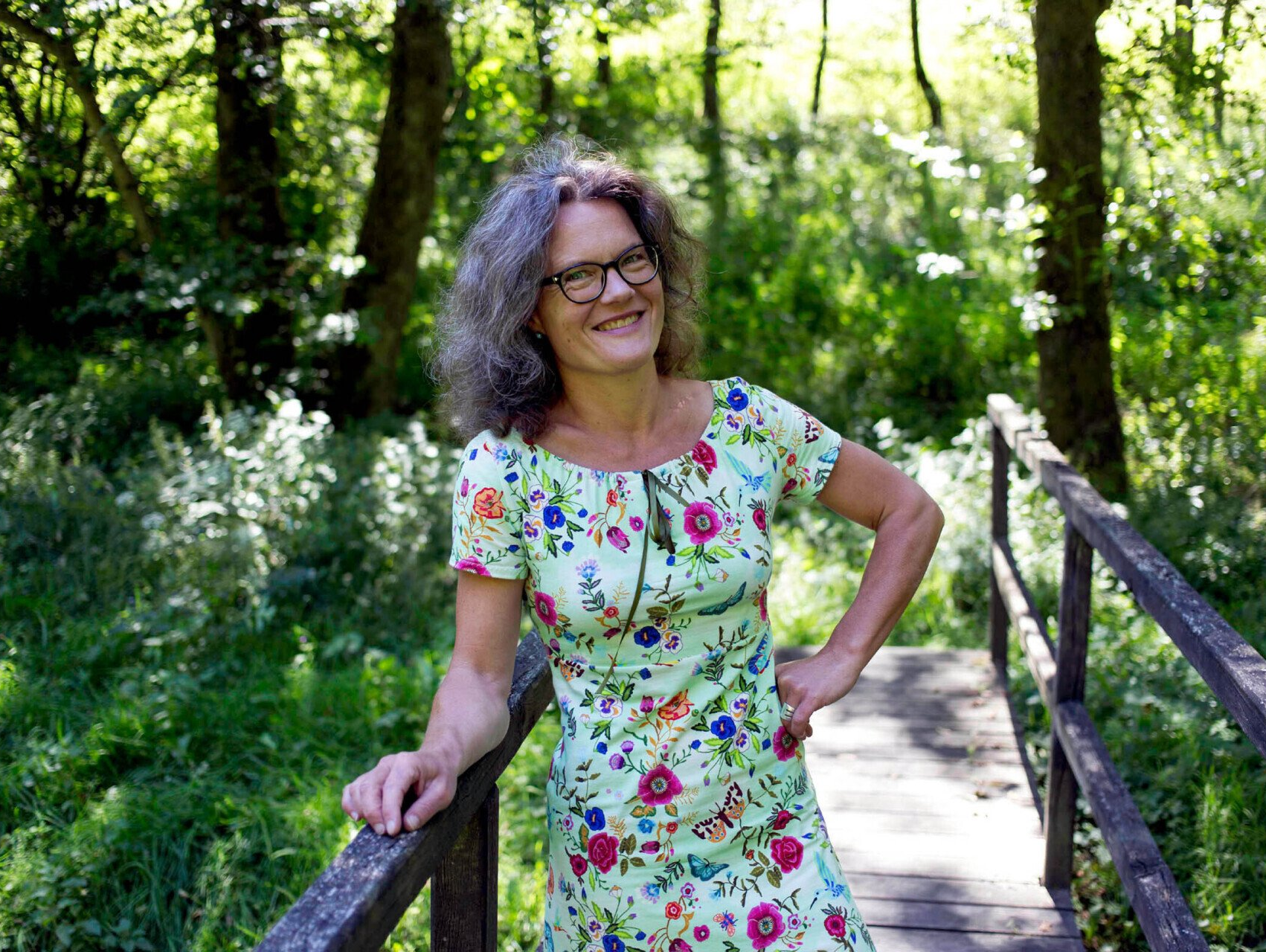 Frau Boecking steht auf einem Steg in einem Bachtal im Grünen und lächelt in die Kamera
