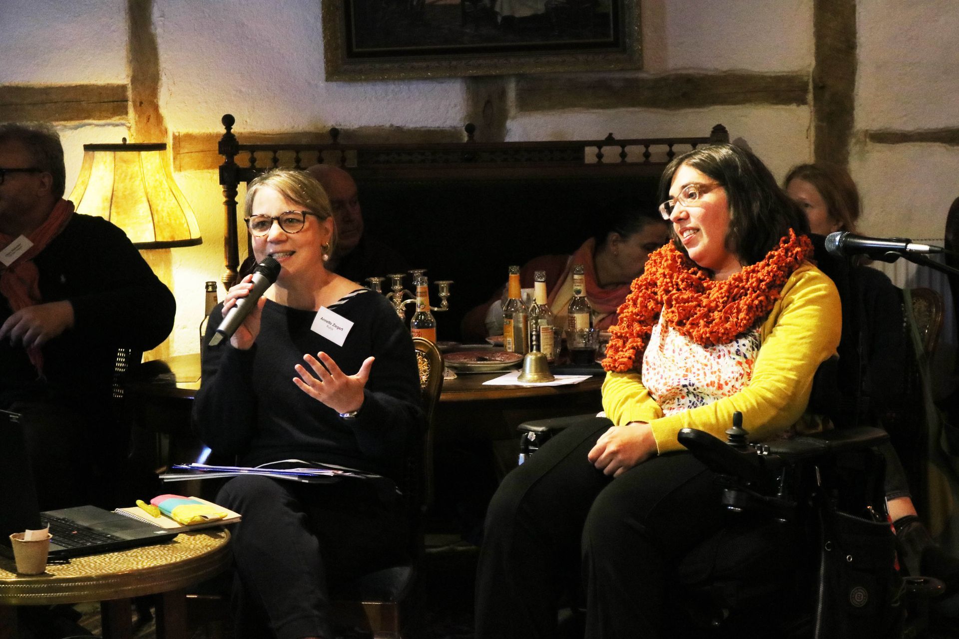 Frau Ziegert und Frau Rosenberg von kubia sitzen an einem Tisch, Frau Ziegert hält das Mikrofon und erzählt. Frau Rosenberg sitzt in einem Rollstuhl.