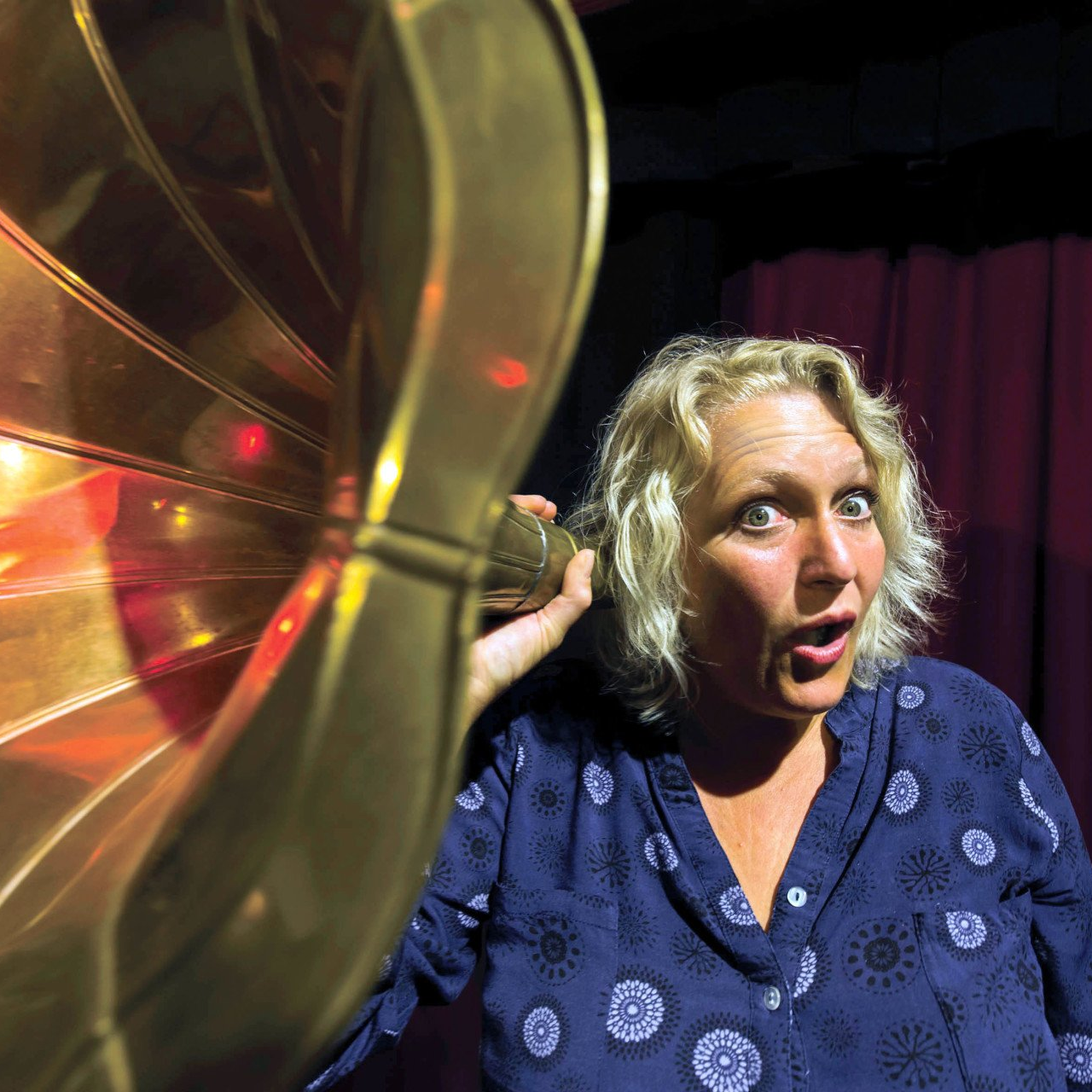 Ulrike Wesely steht vor einem roten Theatervorhand, hält sich einen Grammophontrichter vor ihr rechtes Ohr und macht dabei ein gespielt theatralisch erstauntes Gesicht.
