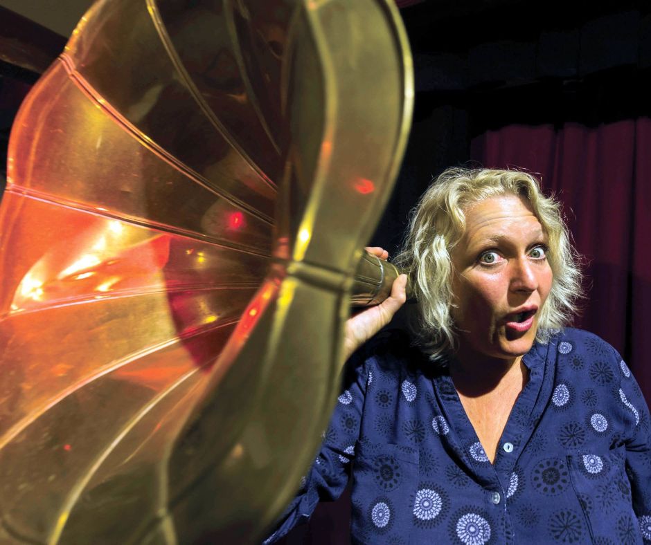 Ulrike Wesely steht vor einem roten Theatervorhand, hält sich einen Grammophontrichter vor ihr rechtes Ohr und macht dabei ein gespielt theatralisch erstauntes Gesicht.