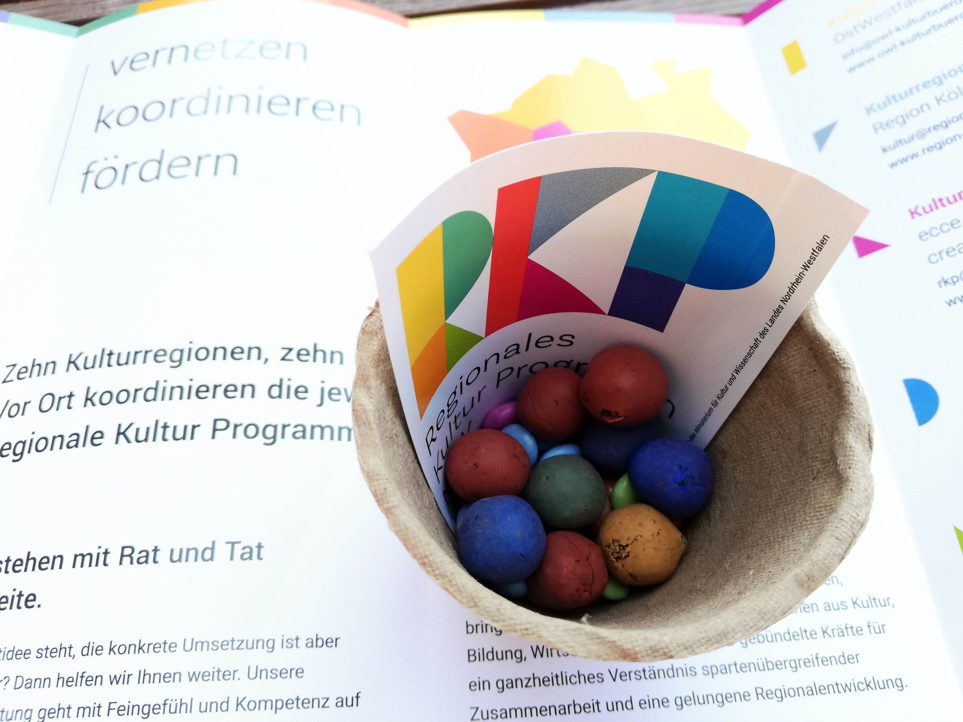 Pflanztopf aus Pappe, darin bunte Blumenmurmeln und Schokolinsen und Aufkleber Regionales Kultur Programm NRW. Im Hintergrund der RKP-Flyer.