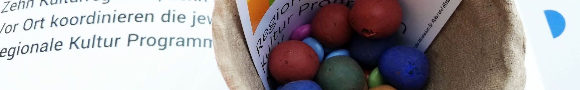 Pflanztopf aus Pappe, darin bunte Blumenmurmeln und Schokolinsen und Aufkleber Regionales Kultur Programm NRW. Im Hintergrund der RKP-Flyer angerissen.