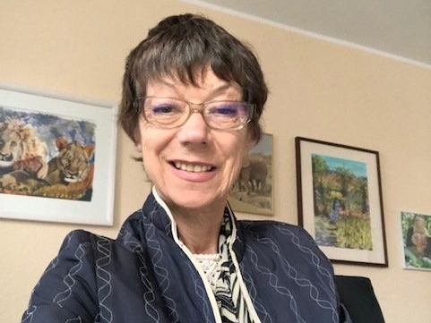 Frau Schneider lächelt in die Kamera, auf der Wand im Hintergrund sind mehrere Gemälde zu sehen