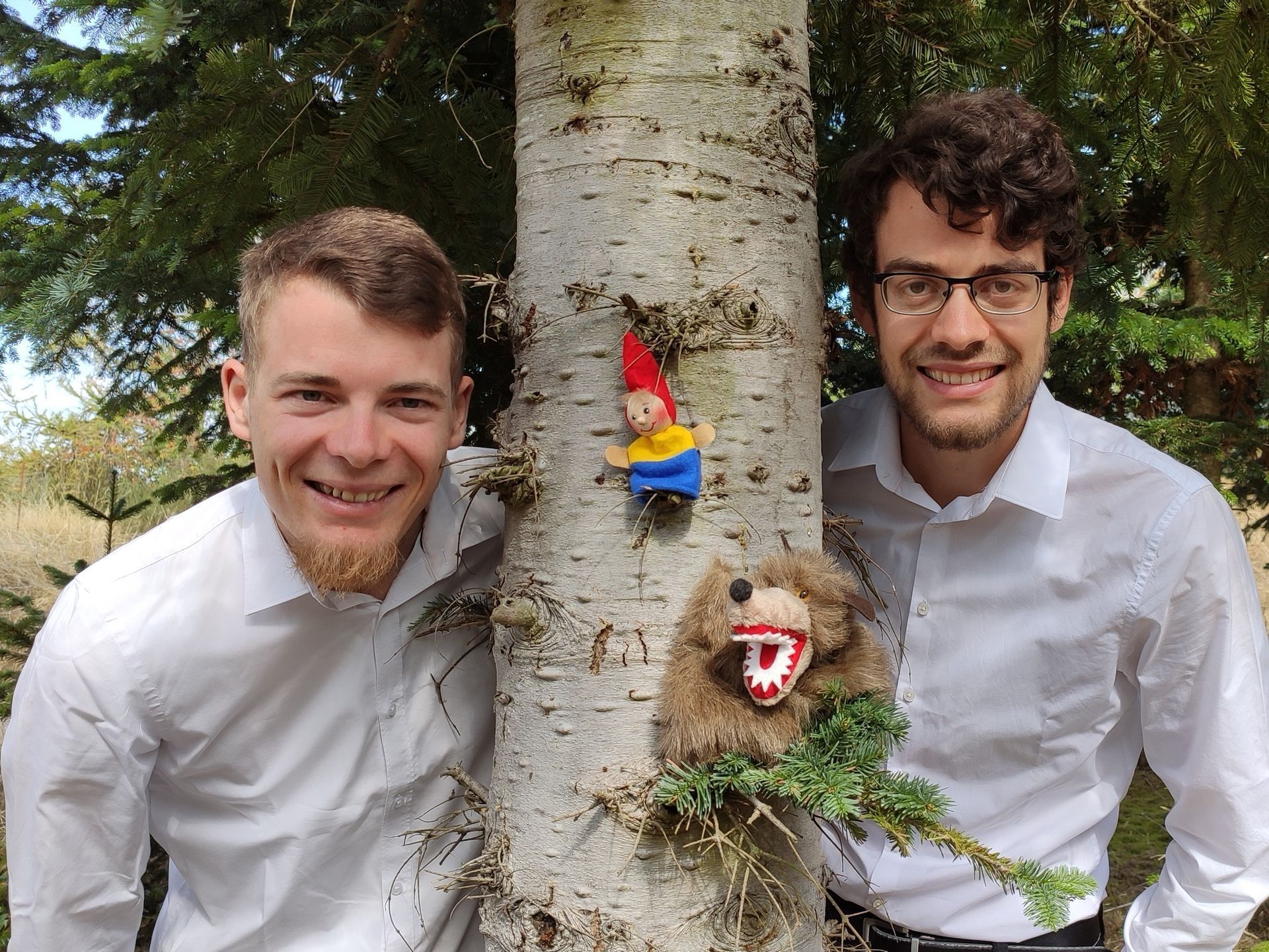 Die Griese-Brüder: zwei junge Männer in weißen Hemden schauen links und rechts hinter einer Fichte hervor. An dem Baumstamm hängt ein kleiner Stoffzwerk und auf einem Ast sitzt ein Kuscheltier (Wolf?)