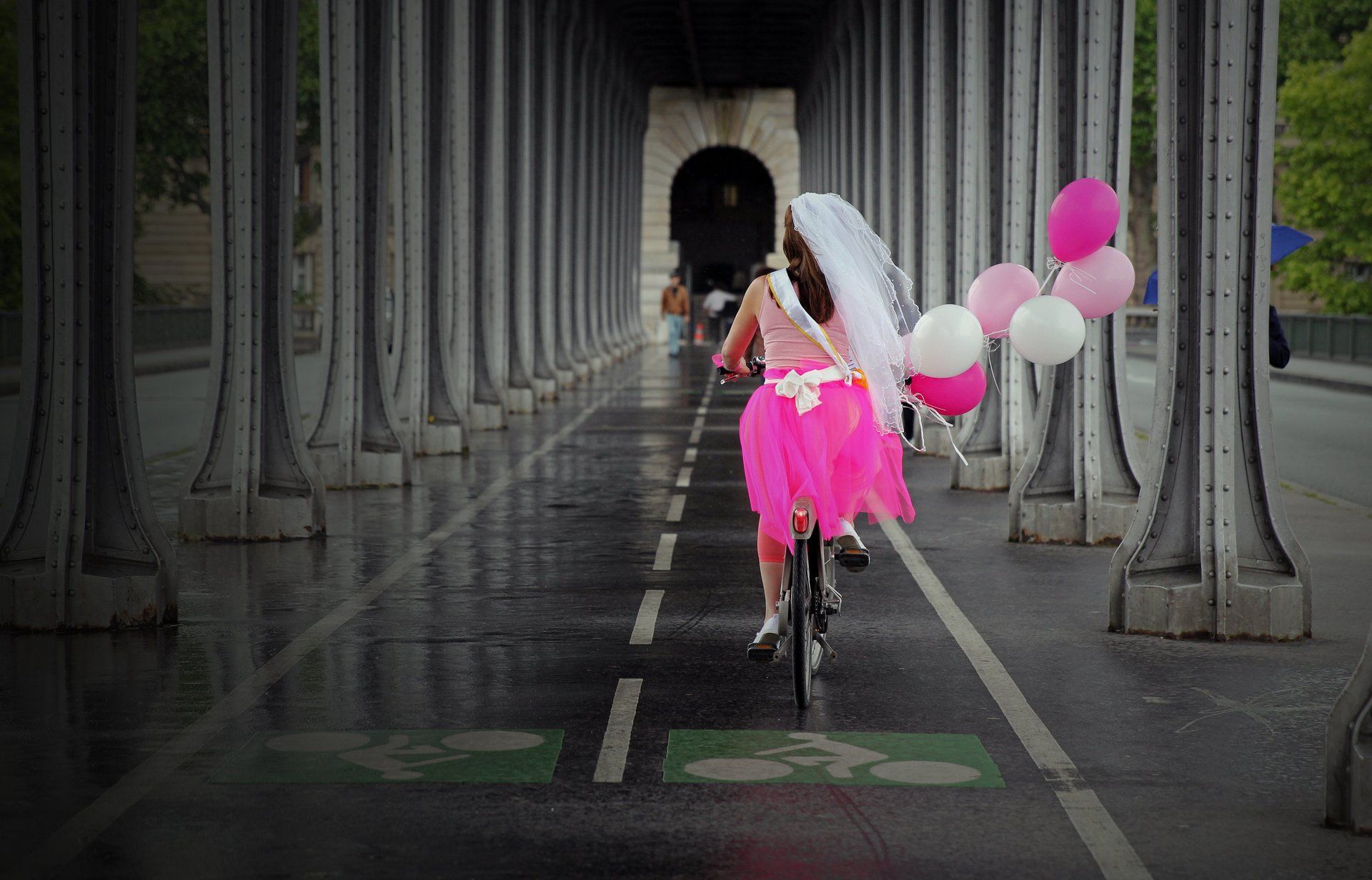 EVJF Jeune fille avec un voile sur la tête, vêtue de rose fuchsia en train de faire du vélo avec des ballons colorés sur une voir de chemin de fer