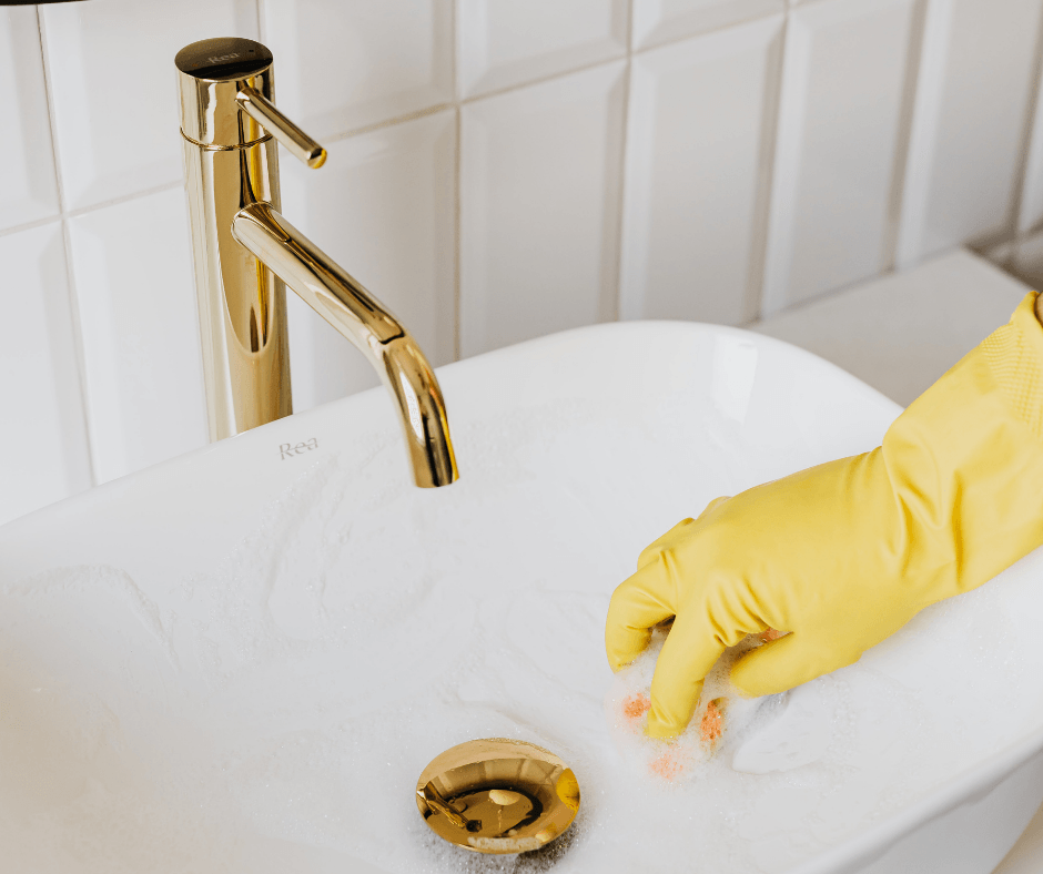 Gloved hand cleaning a white sink with soapy water under a gold tap