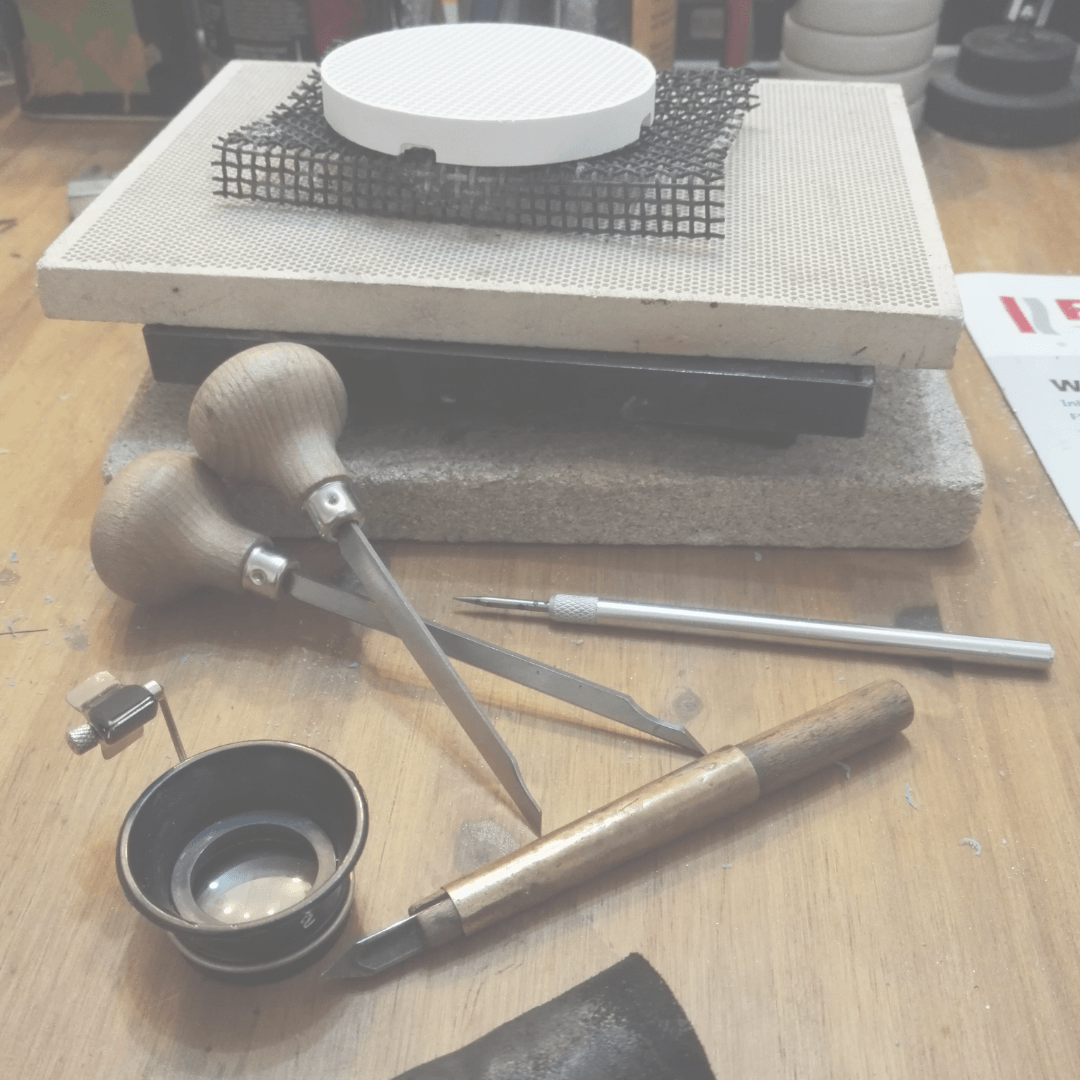 Goldschmiedewerkzeug mit Sticheln, einer Reißnadel, einem Schaber und einer Lupe liegen vor der Lötunterlage