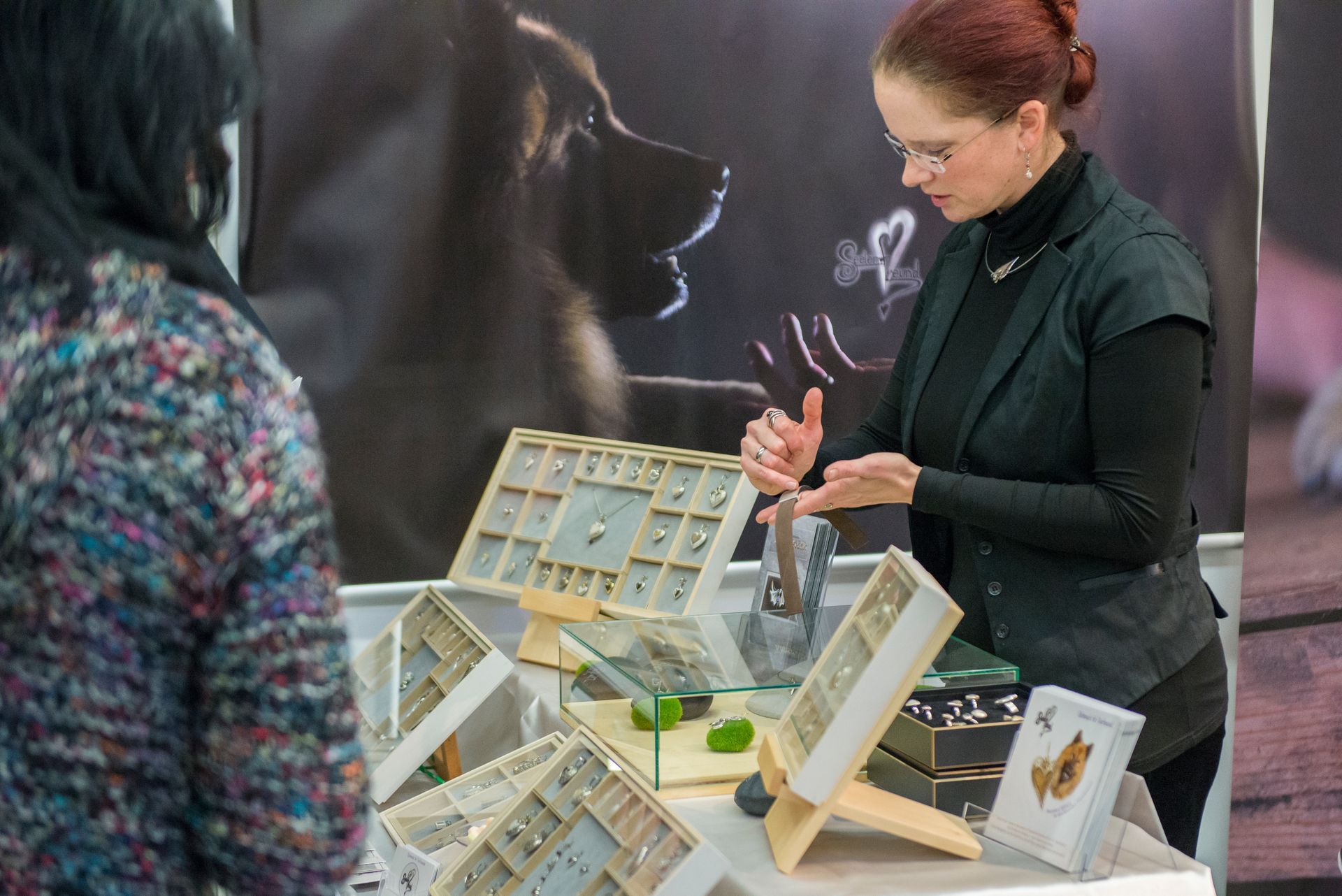 Kundengespräch am Messestand von Seelenfreund auf der Heimtiermesse im Rahmen der Consumenta.
Die Goldschmiedin erklärt die Funtion und zeigt die feinen Details eines Armbandes aus robusten und sehr angenehm zu tragendem Fettleder mit einem Mittelteil aus Silber, in welches die Haare eingefasst werden.
Das Armband ist mit einem Dornverschluss aus Edelstahl versehen und dieses Schmucklstück besticht durch ein schichtes und sehr auffälliges Design.
Seelenfreundschmuck aus Heroldsberg auf der Heimtiermesse in Nürnberg im Jahr 2018