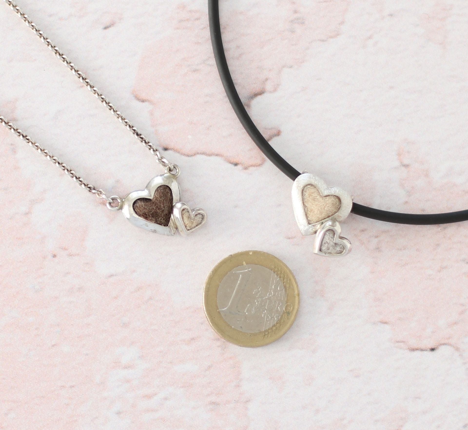 zwei Kettenanhänger mit je zwei Herzen mit Tierhaaren neben einer 1 euro Münze