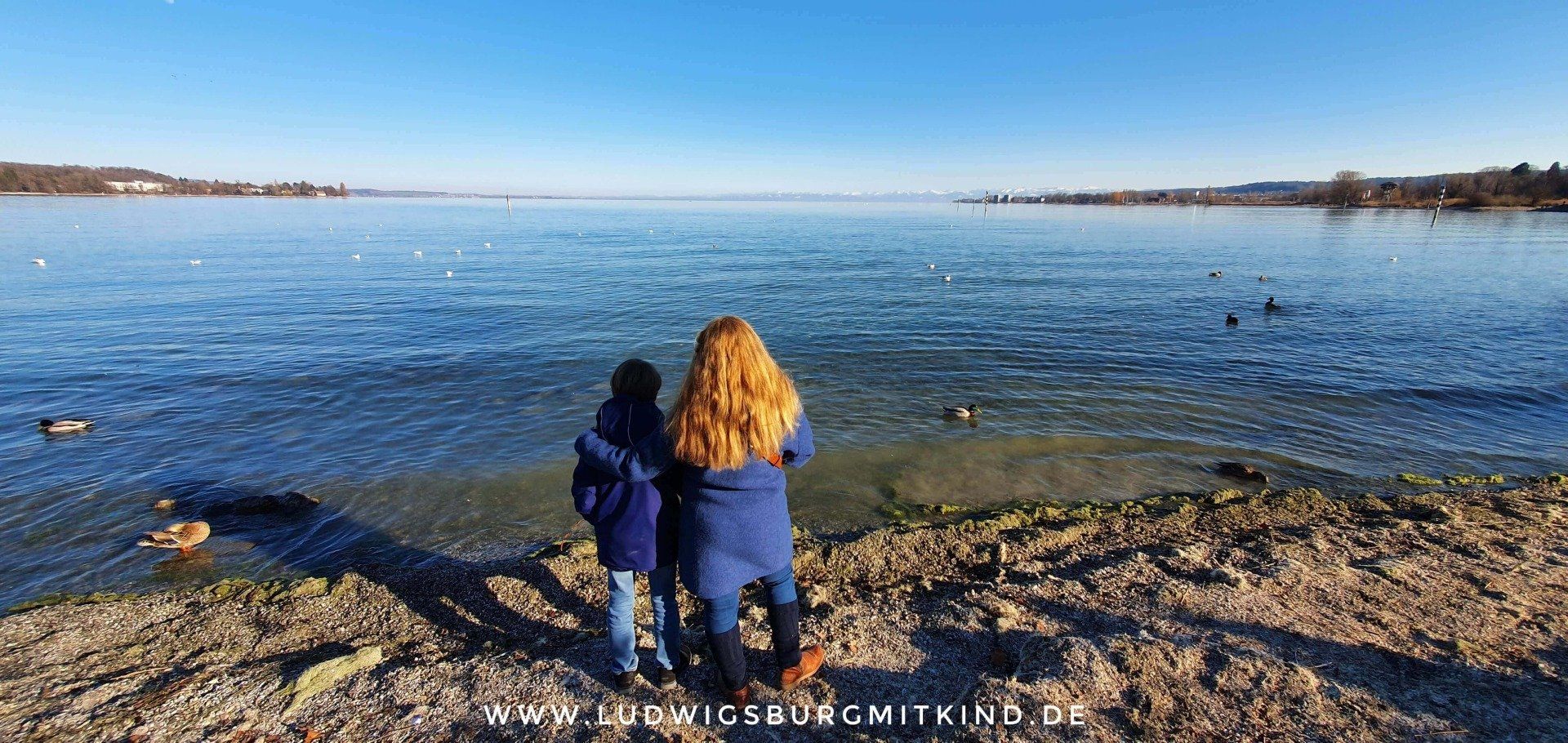 Mama und Sohn stehen am Bodensee und blicken auf den See hinaus.