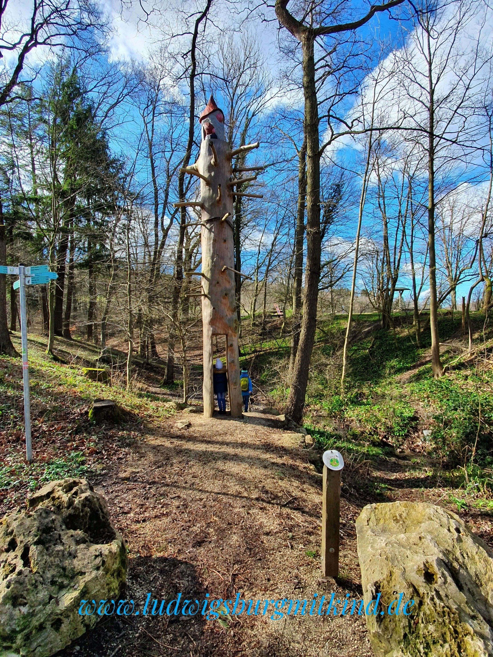 Der Eingang zum Sinneswandel Walderlebnispfad aus einem großen Baum, der zu einem Zweg geschnitzt ist. Durch diesen Eingang gehen Kinder.