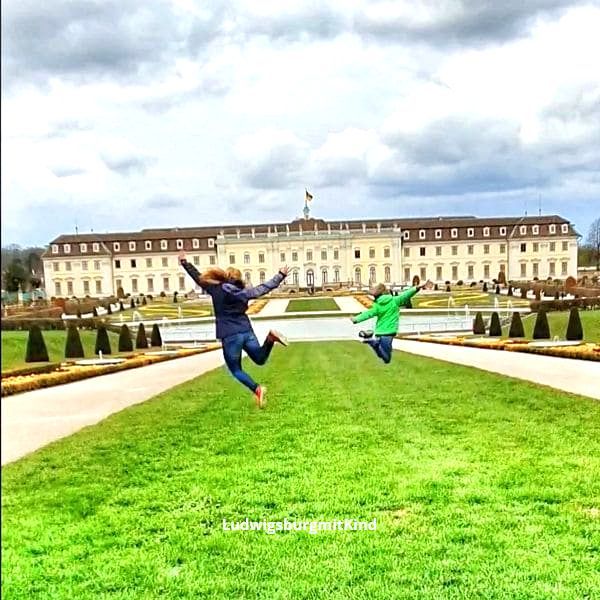 Hier springt eine Mama mit ihrem Kind vor dem Residenzschloss in  Ludwigsburg in die Luft. Das Schloss ist das Wahrzeichen von Ludwigsburg und ein Ausflugsziel für Familien.