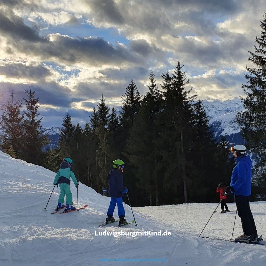 Zwei Kinder stehen mit ihrem Vater auf einer Skipiste, dahinter ist ein Wald und ein spektakuläres Sonne-Wolken-Bild zu sehen.