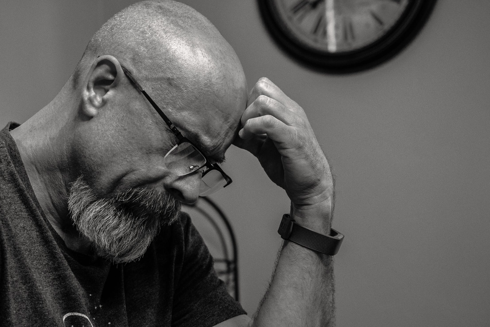 A bald man frowns and looks down at a table, wearing a dark tee-shirt. The picture is in black and white