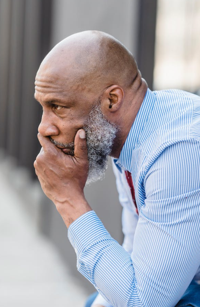 A bald man in a blue shirt looks straight ahead pensively with his chin resting in his hand.