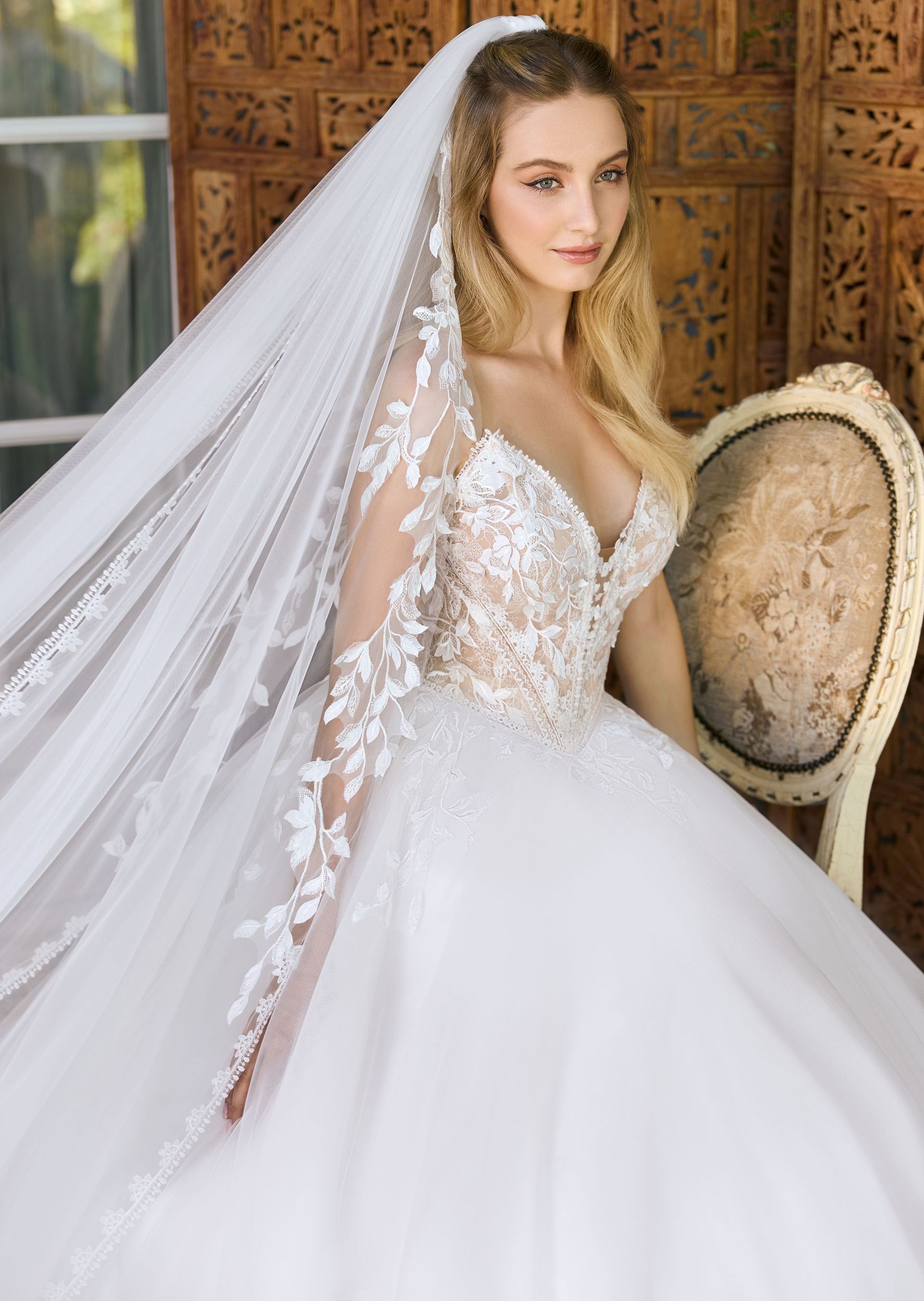 Eine junge Braut trägt ein schulterfreies Brautkleid im Prinzessinnen-Look mit tiefem V-Ausschnitt und v-förmiger Corsage. Das Kleid ist mit feiner Blätterspitze und Blütenspitze verziert. Die Braut trägt einen langen Schleier, der ebenfalls mit filigraner Blätterspitze bestickt ist. Das Brautkleid gehört zur Kollektion von AMORELY Brautmoden Velbert,  in der Nähe von Solingen