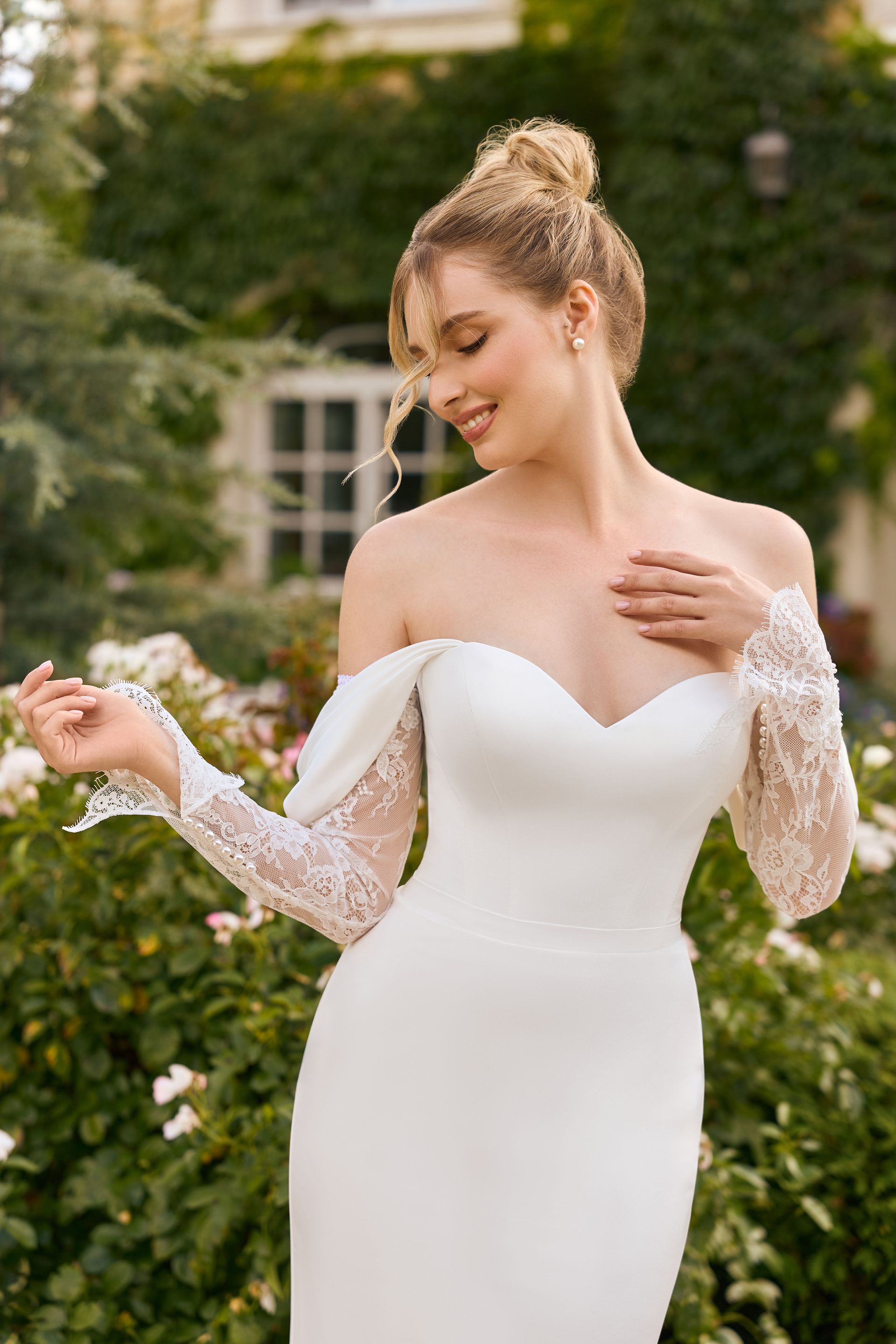 Eine junge Braut trägt ein schulterfreies Fit & Flare-Brautkleid aus glattem Satin mit herzförmigem Ausschnitt und seitlich angesetzten Trägern. Das Kleid hat abnehmbare, lange Ärmel aus französischer Spitze und vereint einen klaren, modernen Look mit romantischen Details. Das Brautkleid gehört zur Kollektion von AMORELY Brautmoden Velbert,  in der Nähe von Oberhausen