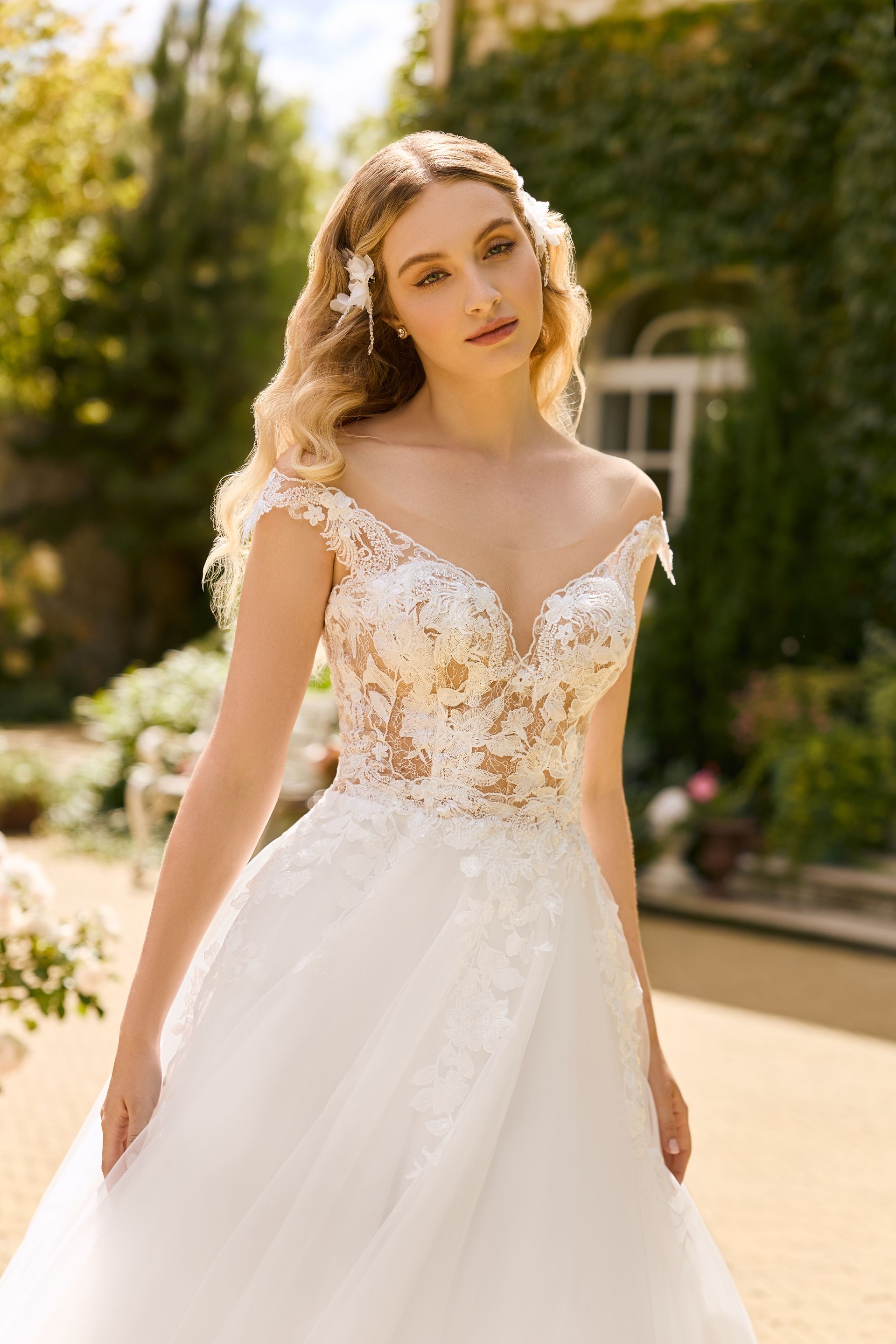 Eine junge Braut trägt ein leichtes A-Linien-Brautkleid aus feinem Tüll. Das transparente Oberteil ist mit einem kleinen V-Ausschnitt gearbeitet und hat schmale, schulterbedeckende Träger, die fließend ins Oberteil übergehen. Das Kleid ist mit zarter Blütenspitze verziert, die über die Taille hinweg in den Rock hineinläuft. Der Look ist luftig, romantisch und ideal für eine Sommerhochzeit. Das Brautkleid gehört zur Kollektion von AMORELY Brautmoden Velbert,  in der Nähe von Dortmund