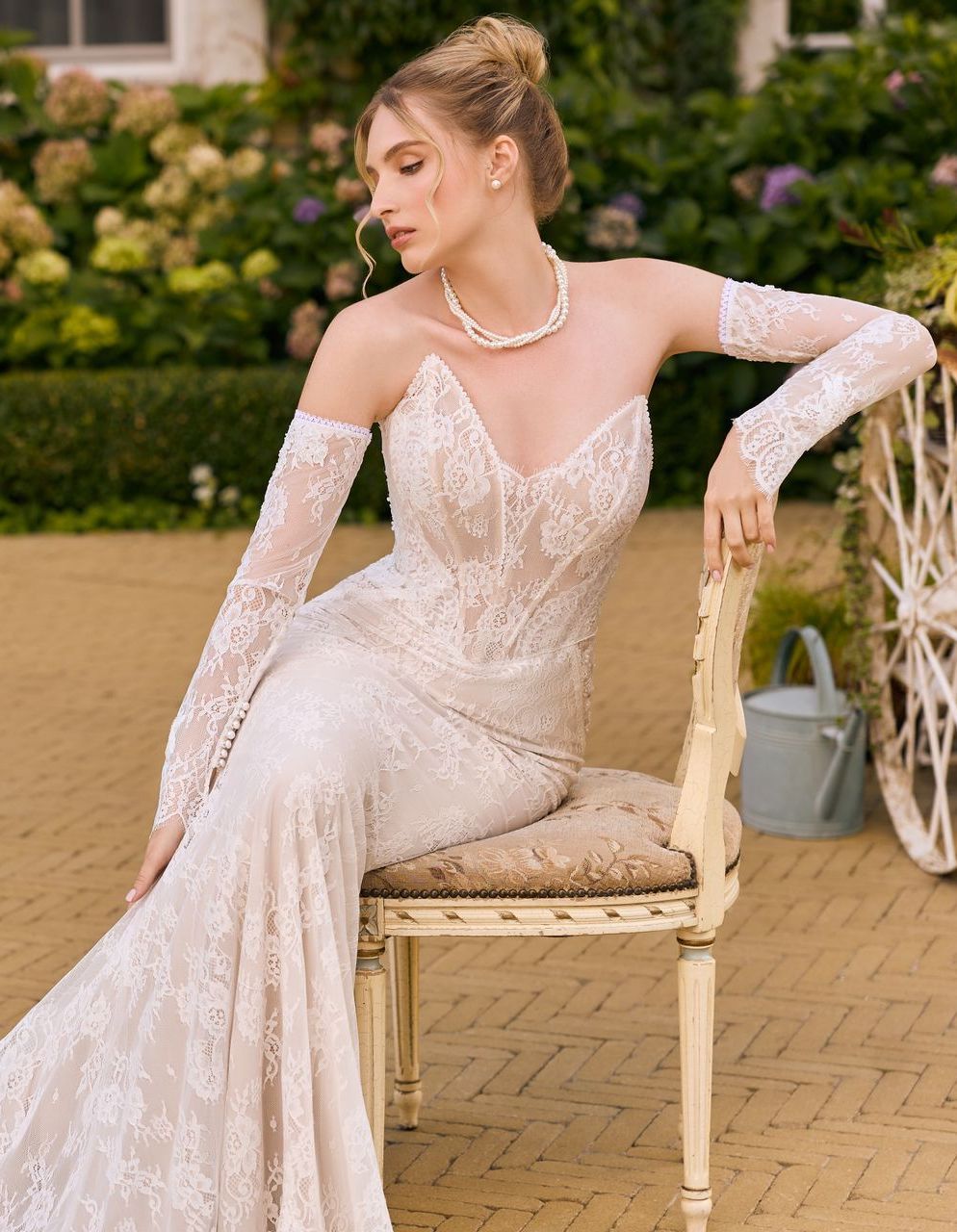 Eine junge Braut trägt ein Fit & Flare-Brautkleid mit v-förmiger Corsage, tiefem V-Ausschnitt und Herzausschnitt. Das Kleid ist komplett aus feiner französischer Spitze gearbeitet. Die schulterfreien Spitzenärmel sind abnehmbar. Die Braut sitzt auf einem Stuhl – das Bild zeigt, dass das Kleid auch im Sitzen bequem ist und die Figur elegant zur Geltung bringt. Das Brautkleid gehört zur Kollektion von AMORELY Brautmoden Velbert,  in der Nähe von Solingen