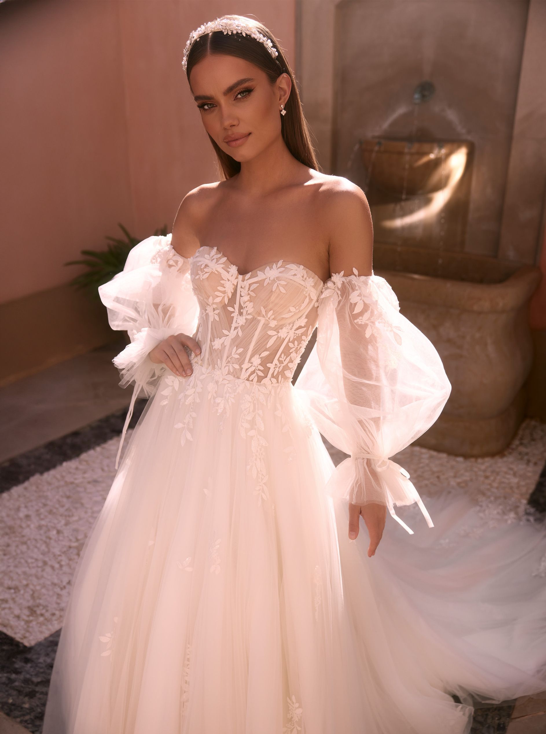 Eine junge Braut trägt ein schulterfreies A-Linien-Brautkleid mit Lingerie-Effekt und transparenter Corsage aus Blütenspitze und 3D-Applikationen. Die abnehmbaren Ballonärmel aus feinem Tüll sind am Handgelenk mit Schleifen gebunden. Der Rock besteht aus mehreren Lagen weichem Tüll und ist mit wunderschöner Blüten-Blätter-Spitze verziert. Das Brautkleid gehört zur Kollektion von AMORELY Brautmoden Velbert,  in der Nähe von  Hilden