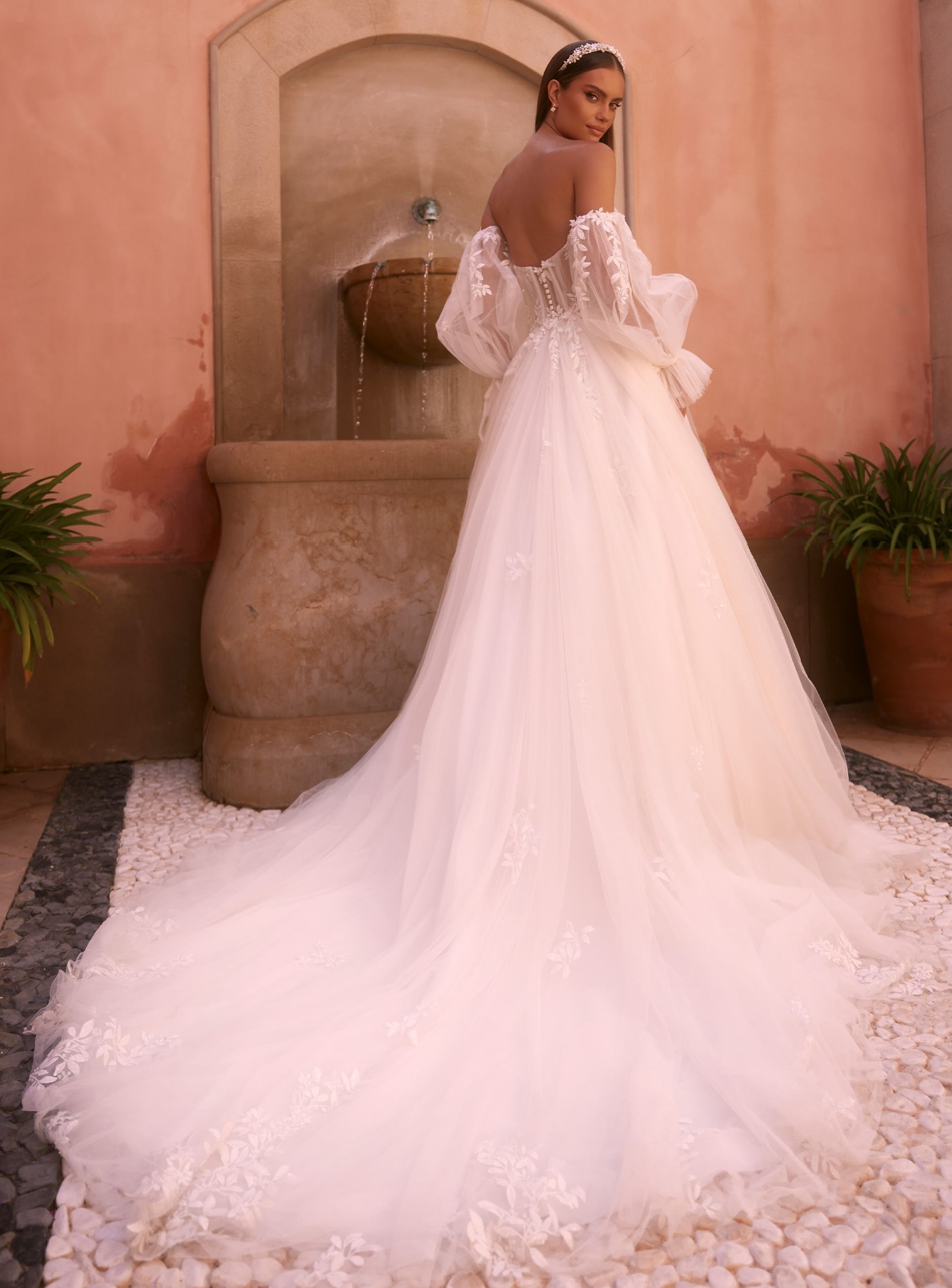 Eine junge Braut trägt ein schulterfreies A-Linien-Brautkleid mit tiefem Rückenausschnitt, transparenter Korsage und abnehmbaren Ballonärmeln aus feinem Tüll. Die lange Schleppe ist mit filigraner Blüten- und Blätterspitze verziert. Das Brautkleid gehört zur Kollektion von AMORELY Brautmoden Velbert,  in der Nähe von Oberhausen