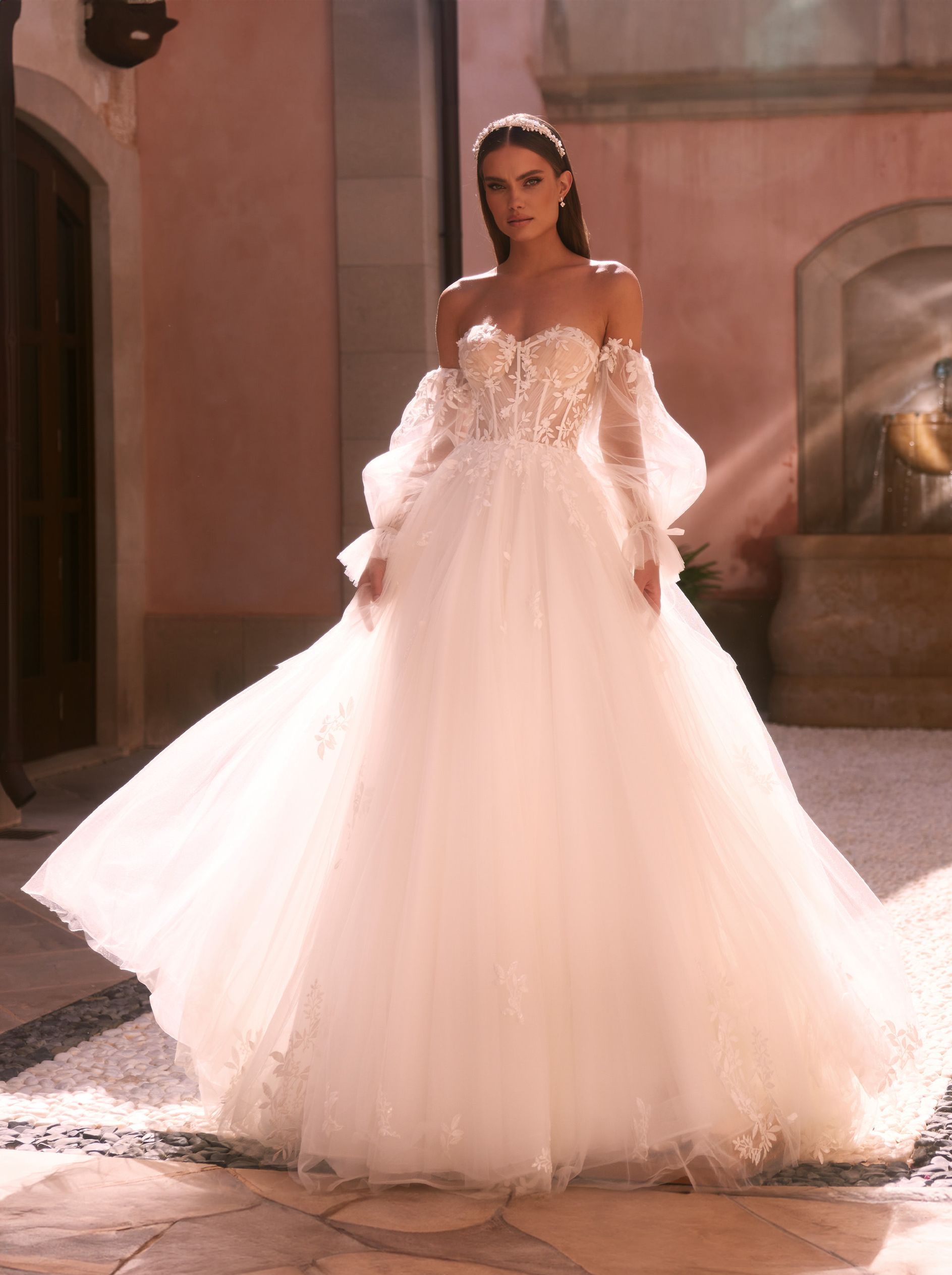 Eine junge Braut trägt ein schulterfreies A-Linien-Brautkleid mit transparenter Corsage im Lingerie-Stil, verziert mit Blütenspitze und 3D-Blüten. Die Ballonärmel aus feinem Tüll sind abnehmbar und mit Schleifen am Handgelenk gebunden. Der mehrlagige Tüllrock ist weit ausgestellt und mit Blüten-Blätter-Spitze dekoriert. Das Brautkleid gehört zur Kollektion von AMORELY Brautmoden Velbert und Wuppertal.
