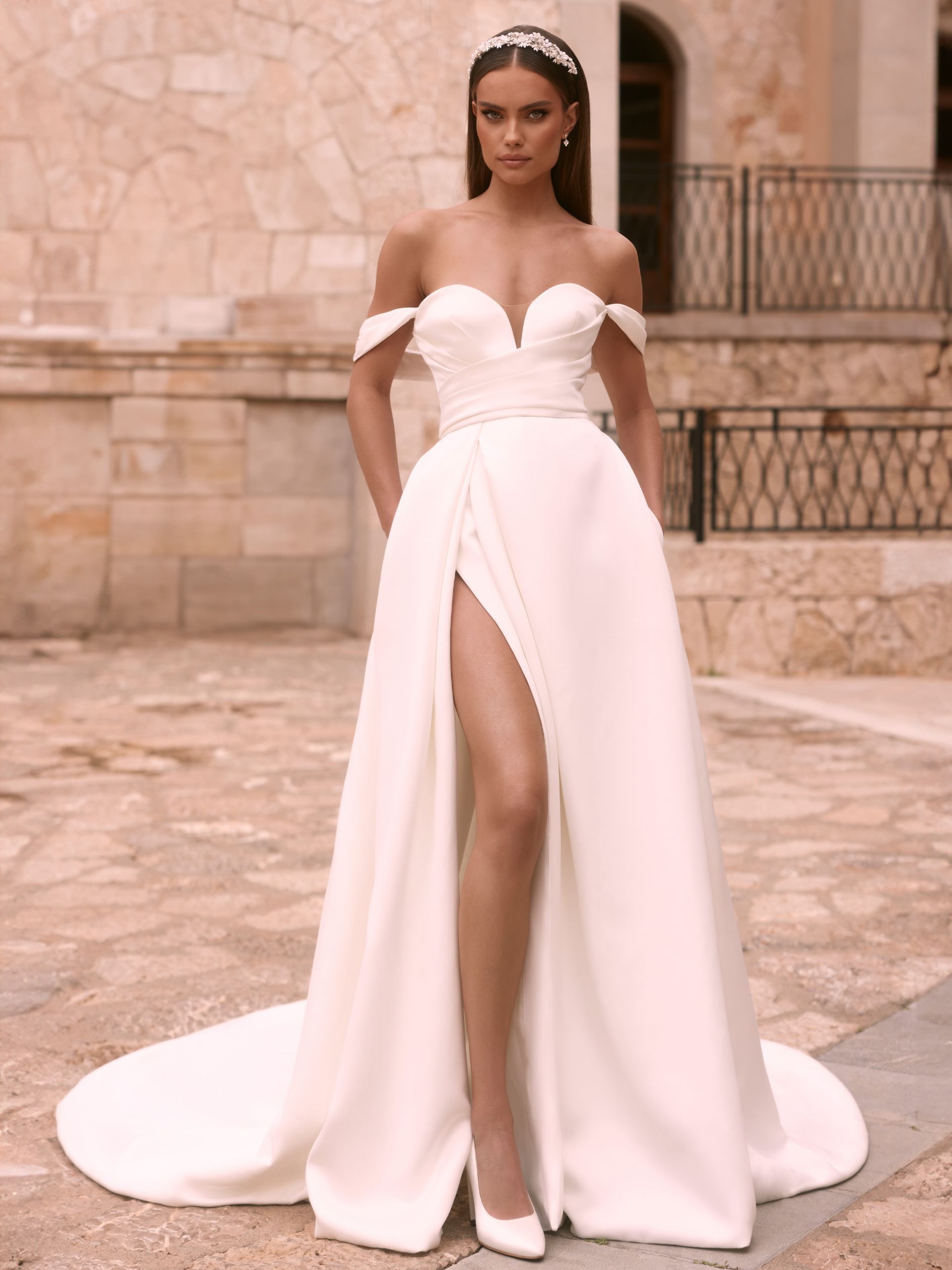 Eine junge Braut trägt ein modernes, schulterfreies Brautkleid im Clean Look aus glattem Satin. Das Kleid hat einen tiefen V-Ausschnitt, seitlich angesetzte Träger und ein asymmetrisch geschnittenes, raffiniertes Oberteil. Der Rock ist ausgestellt, hat einen hohen Beinschlitz und seitliche Eingrifftaschen. Das Brautkleid gehört zur Kollektion von AMORELY Brautmoden Velbert und Wuppertal.
